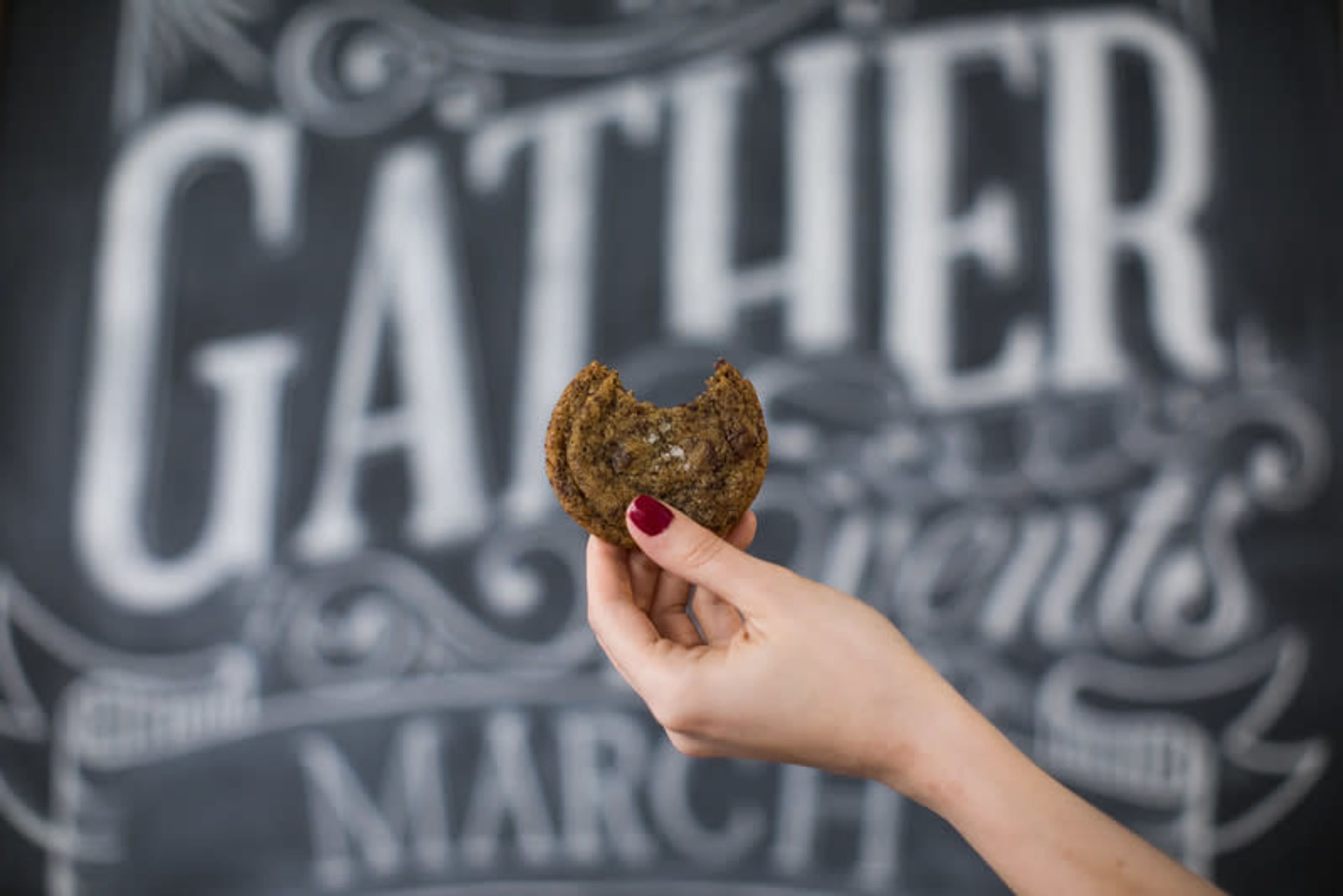 A hand holds a partially eaten cookie in front of a decorative background featuring the word "GATHER."
