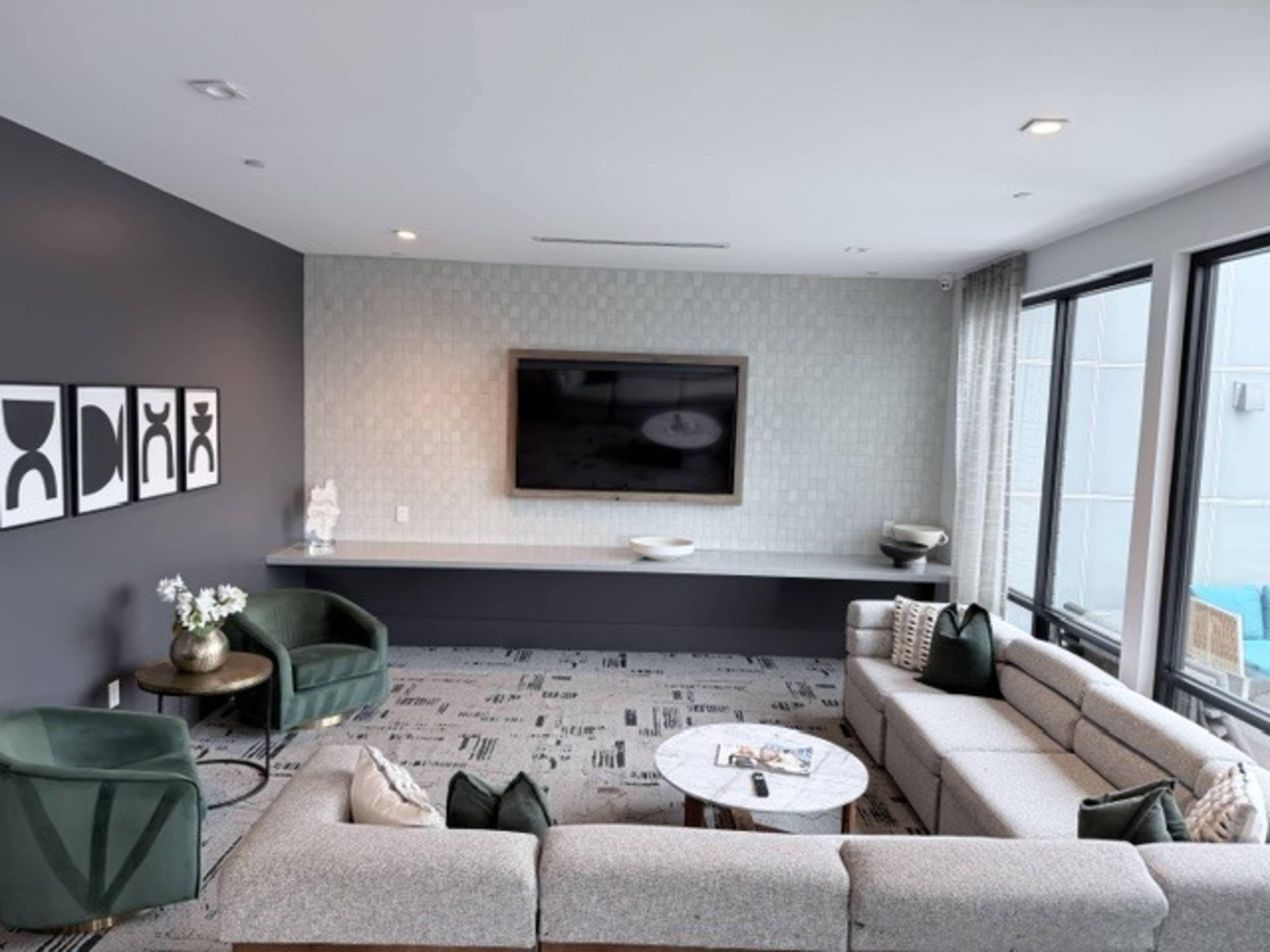The image shows a modern living room featuring a large sectional sofa, a round coffee table, and a wall-mounted television, with framed artwork and large windows.
