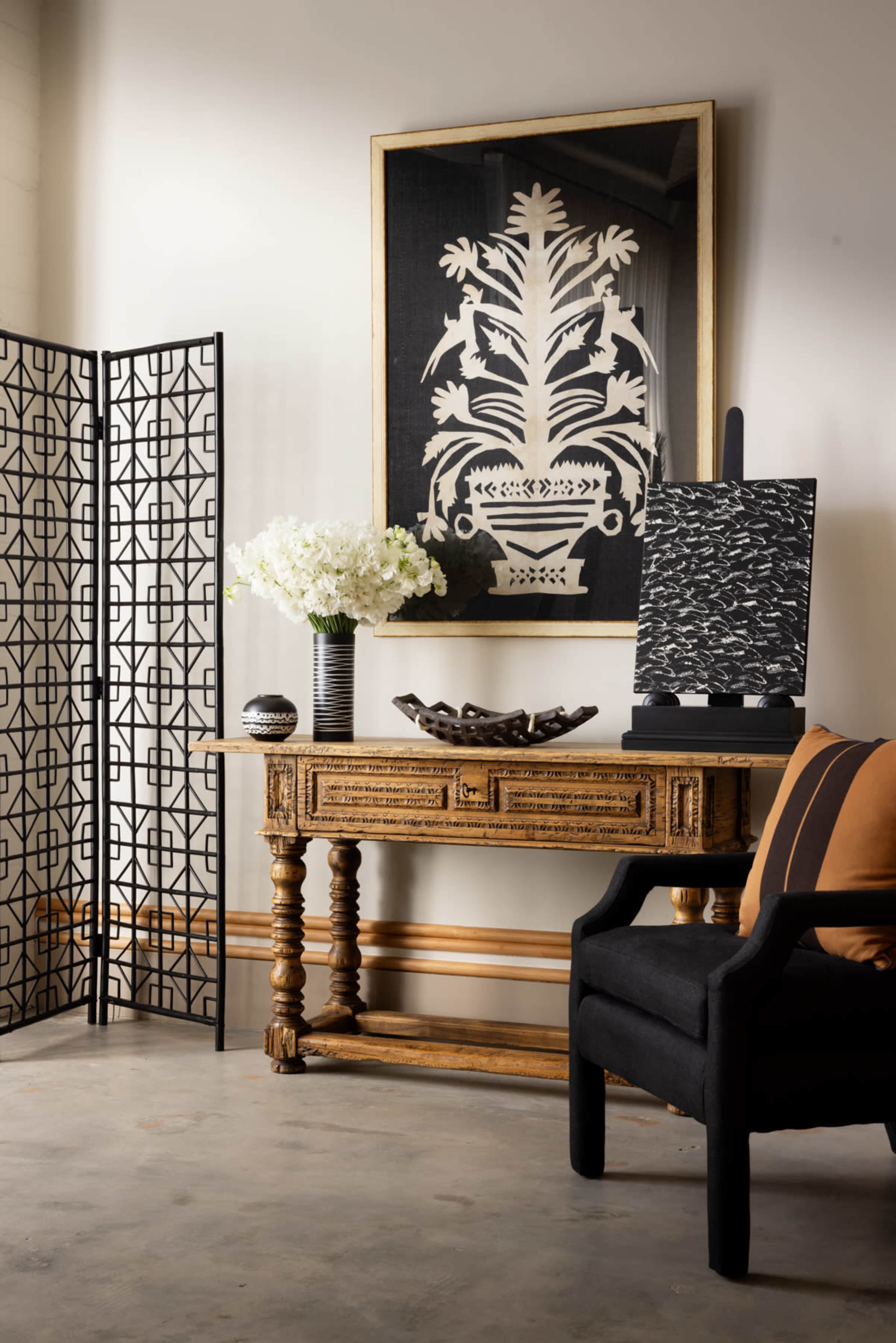 The scene features a wooden console table with a decorative vase of white flowers, a framed artwork on the wall, and a black chair beside a folding screen.