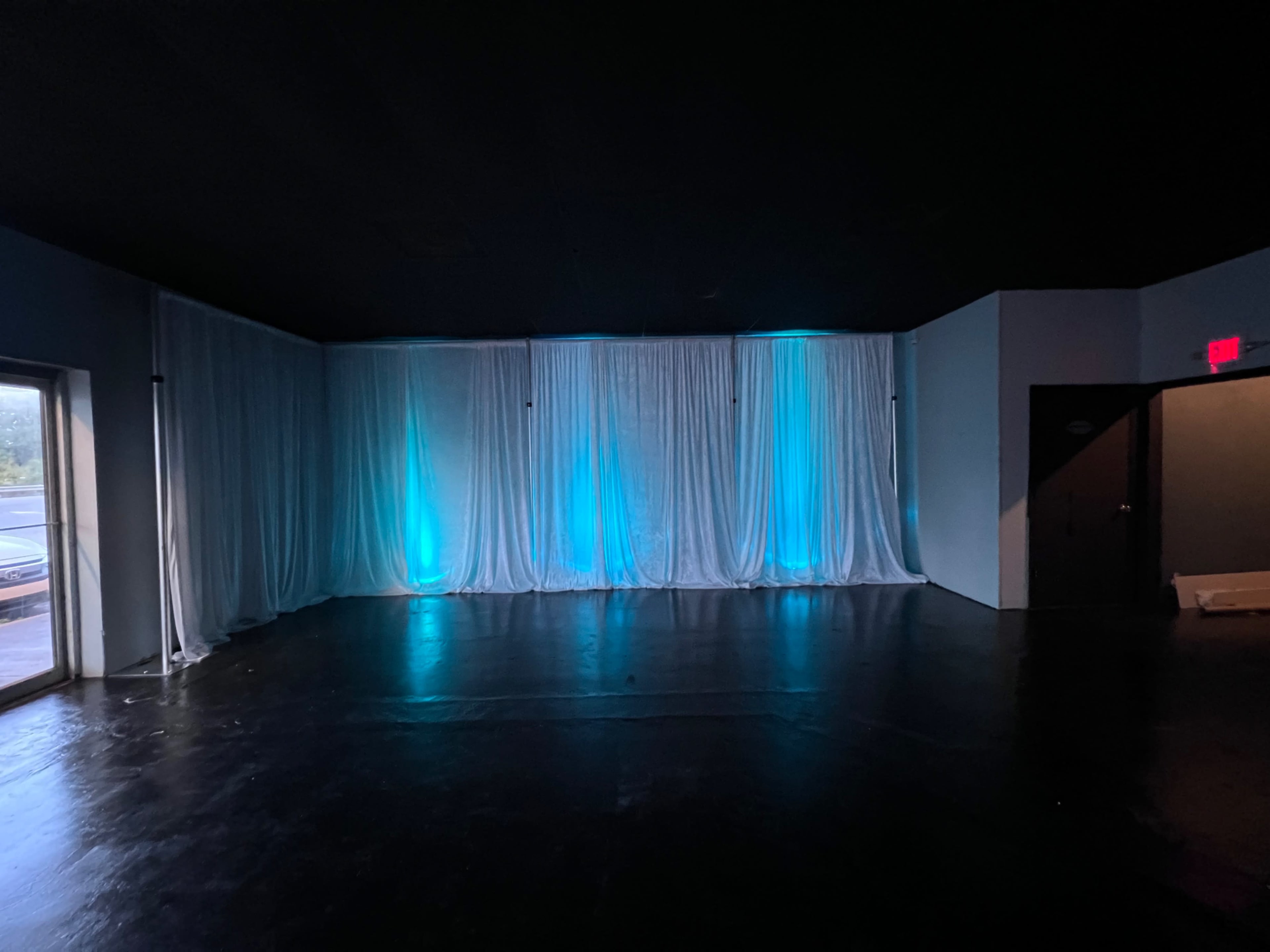 The room is dimly lit with textured black flooring and sheer white curtains backlit in blue along the walls.