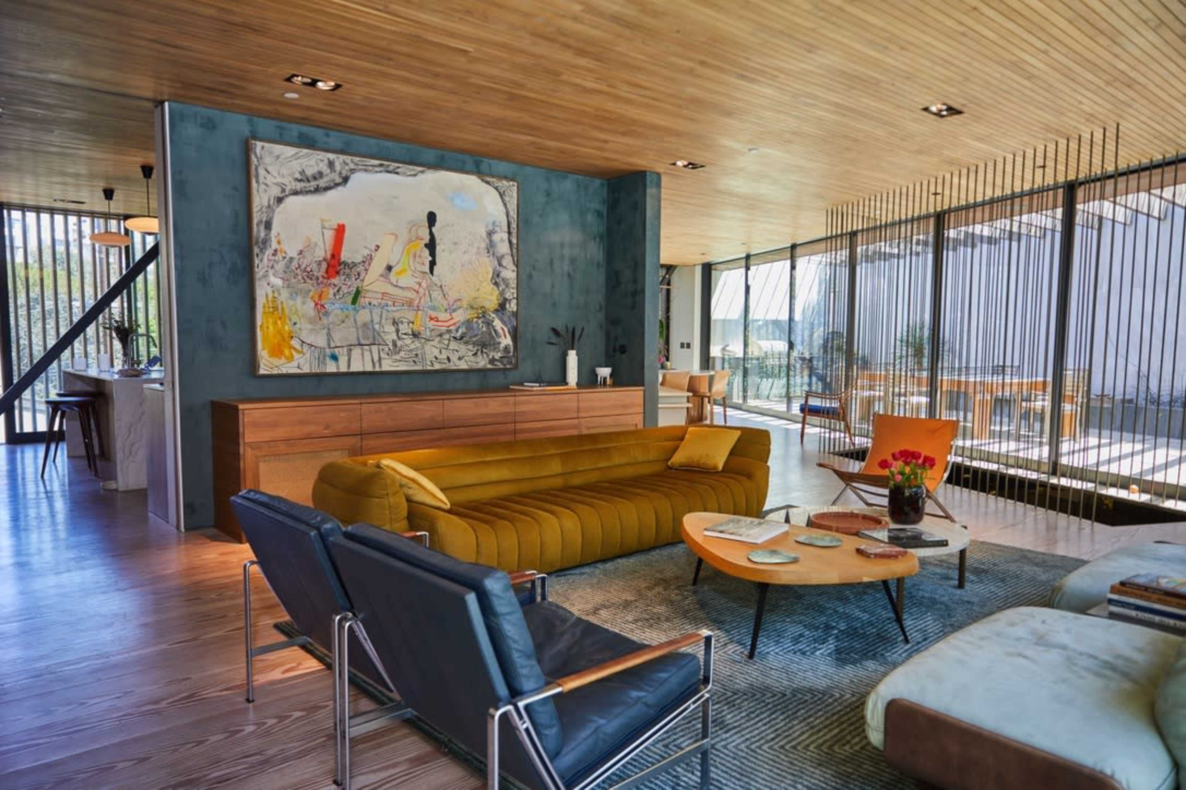 The image shows a modern living room with a yellow sofa, blue armchairs, a wooden coffee table, and large windows that provide natural light.
