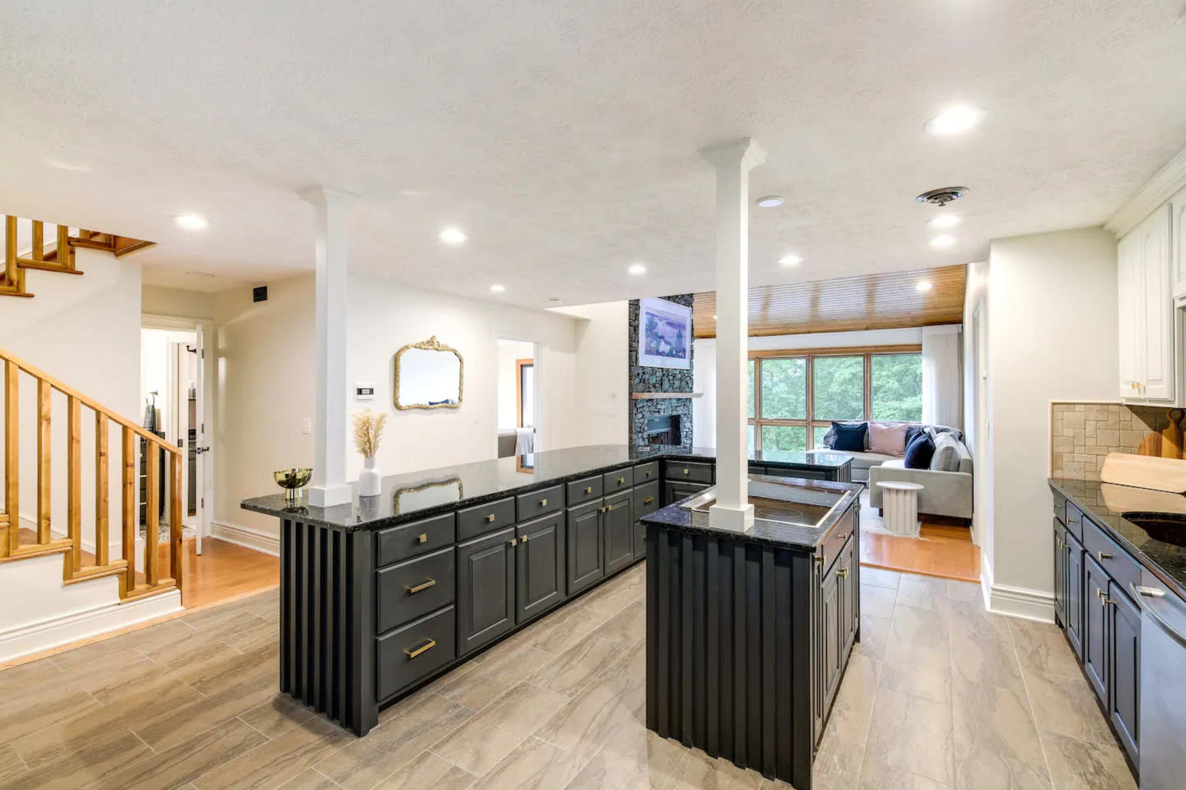 The kitchen features dark cabinetry with gold hardware, a large island, and an open layout that connects to a living area with large windows.