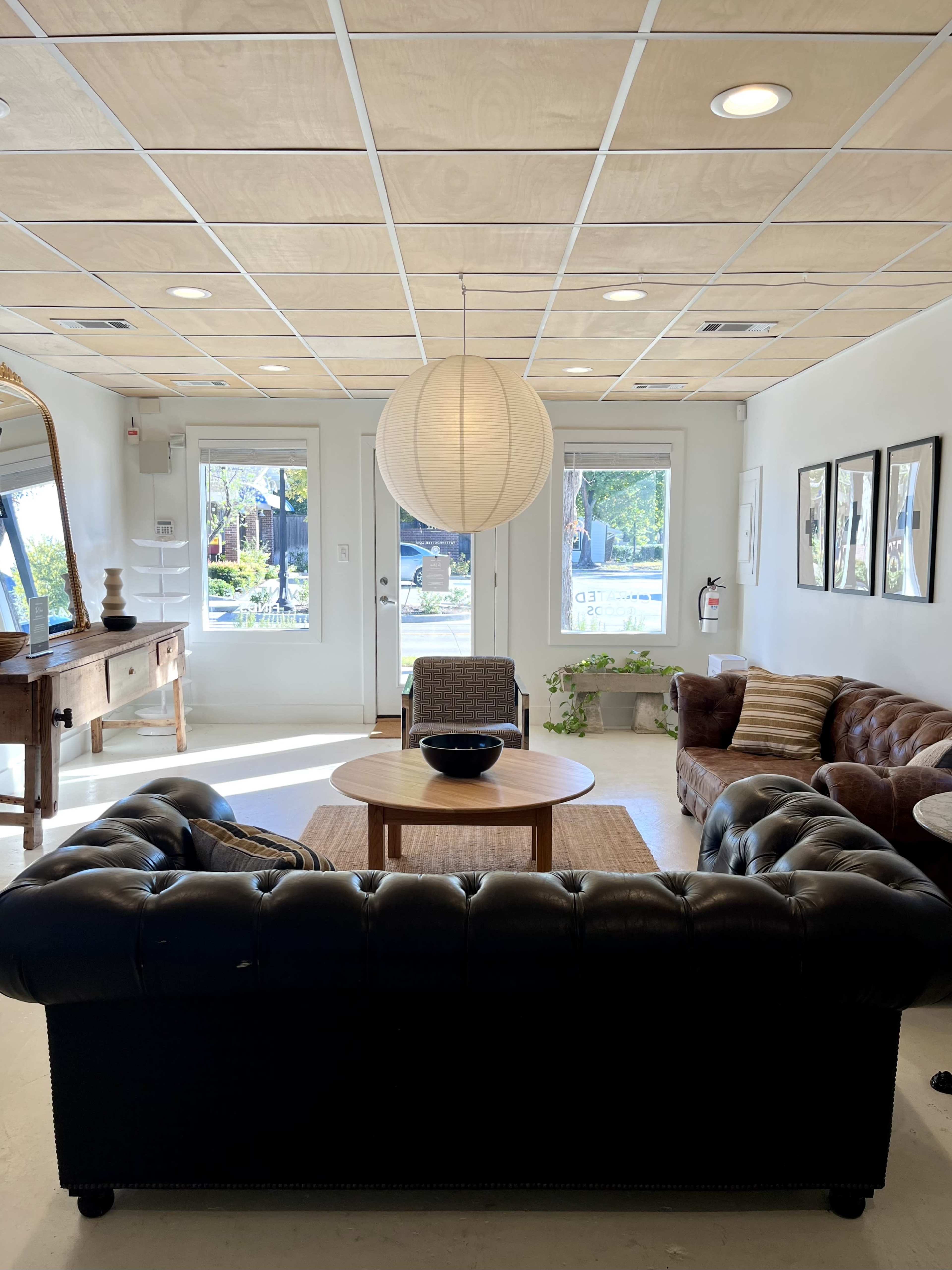 The image shows a modern lounge area with a black leather sofa, a round wooden table, and natural light coming through large windows.
