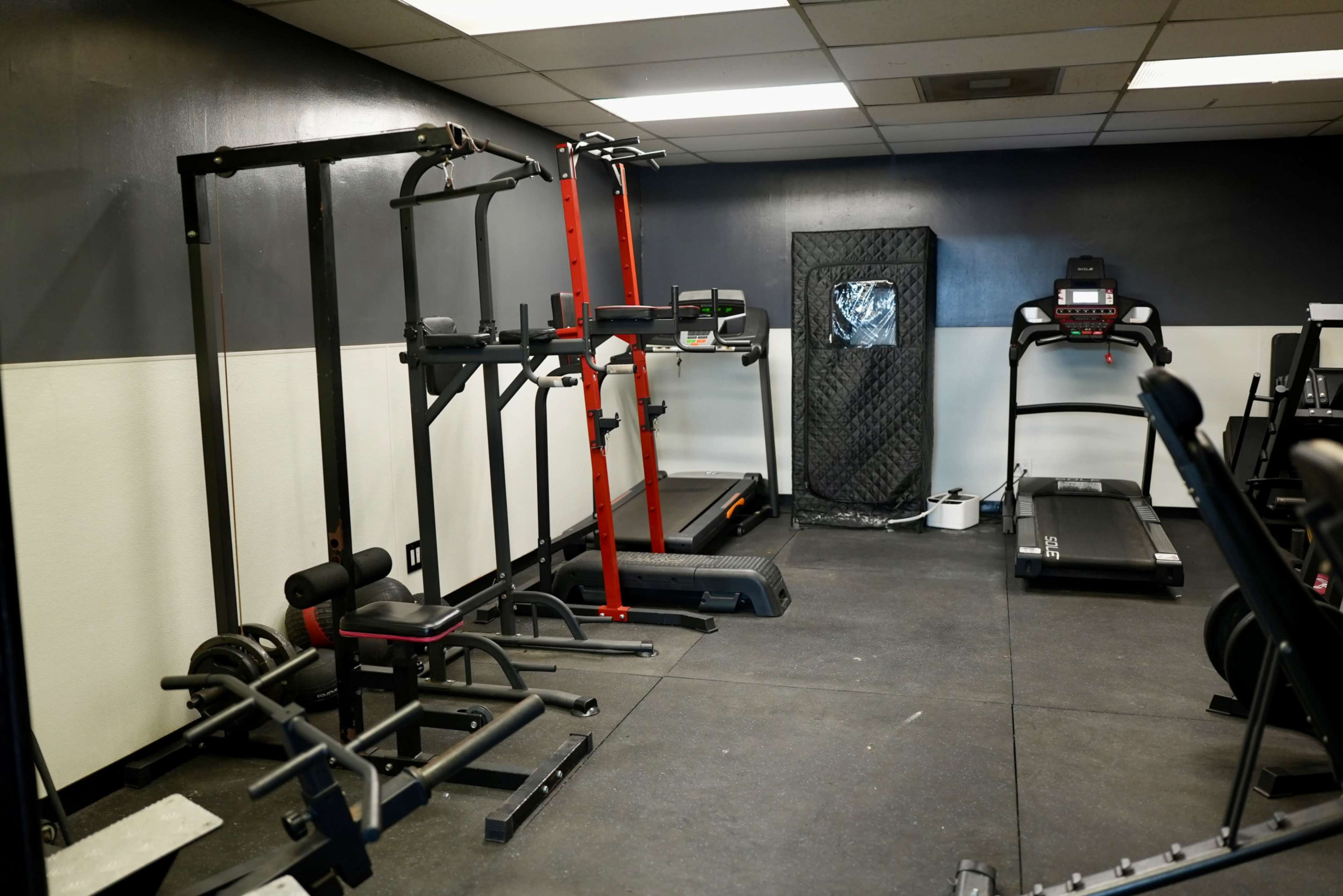 The image shows a small gym area equipped with various fitness machines and equipment, including a treadmill, pull-up bar, and weight rack.