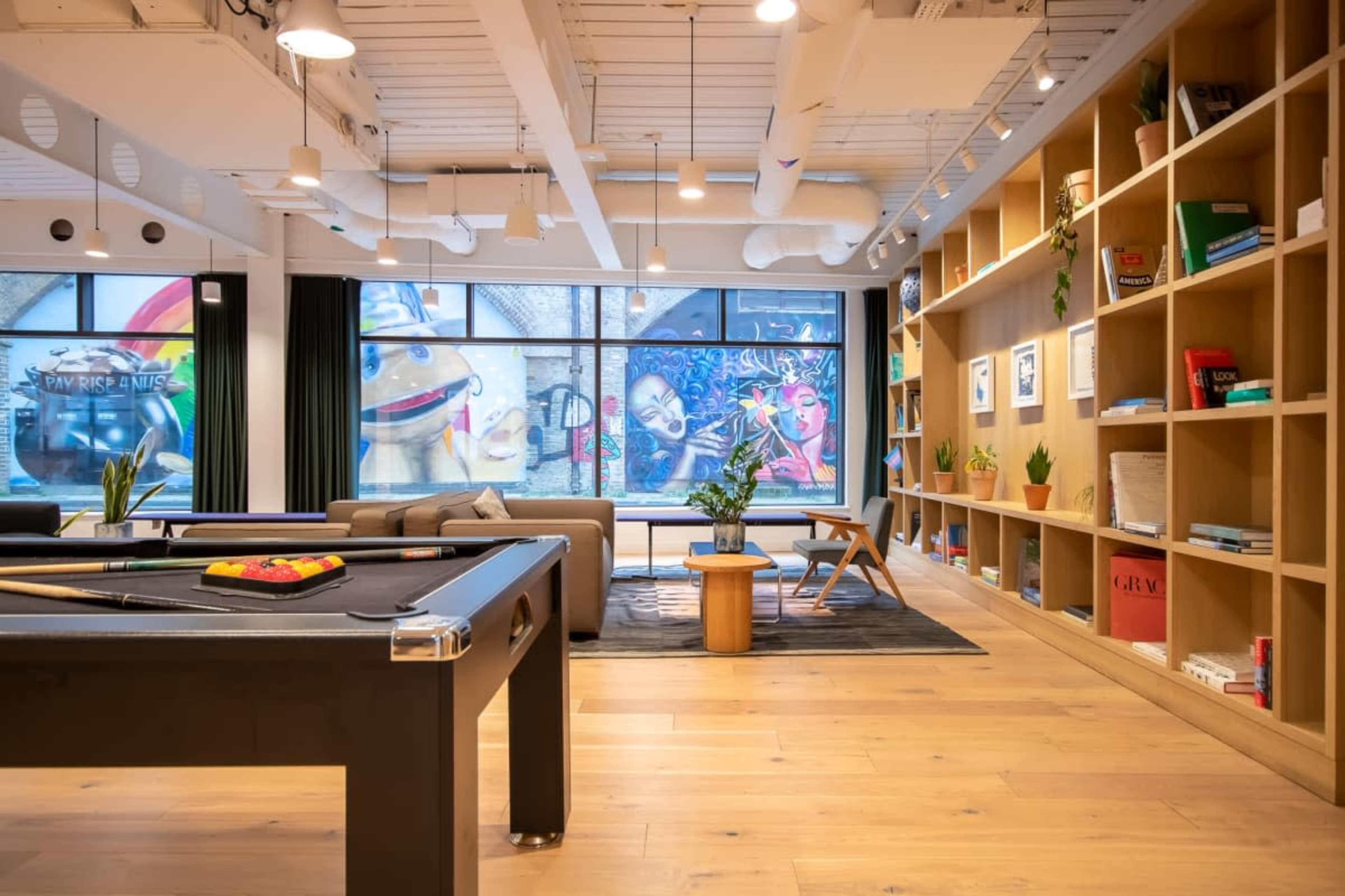 The interior of a modern common area features a pool table, comfortable seating, and a large bookshelf, with a vibrant mural visible through the windows.
