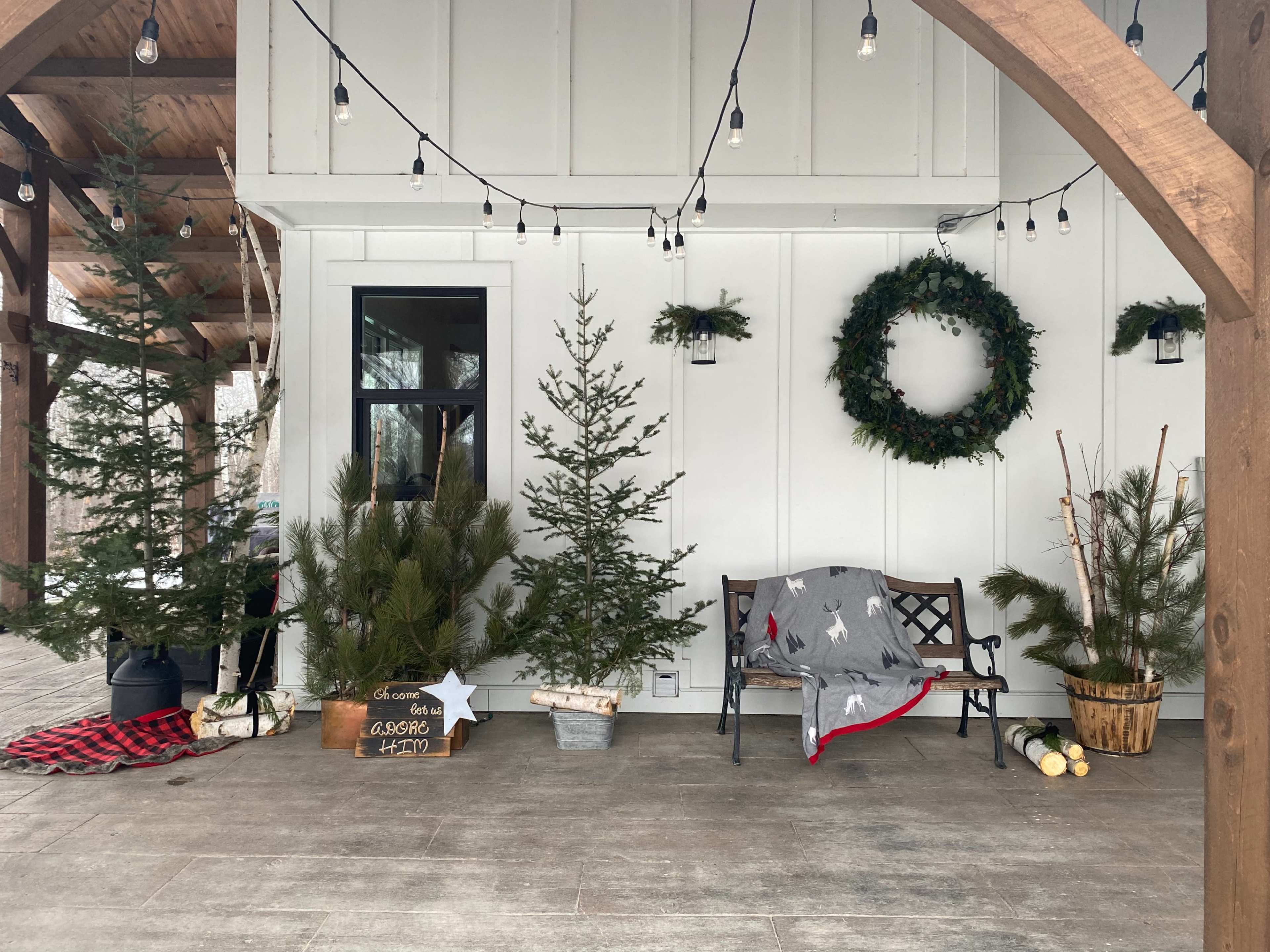 A cozy porch decorated with evergreen trees, a bench, a blanket, and holiday-themed decor, illuminated by string lights.