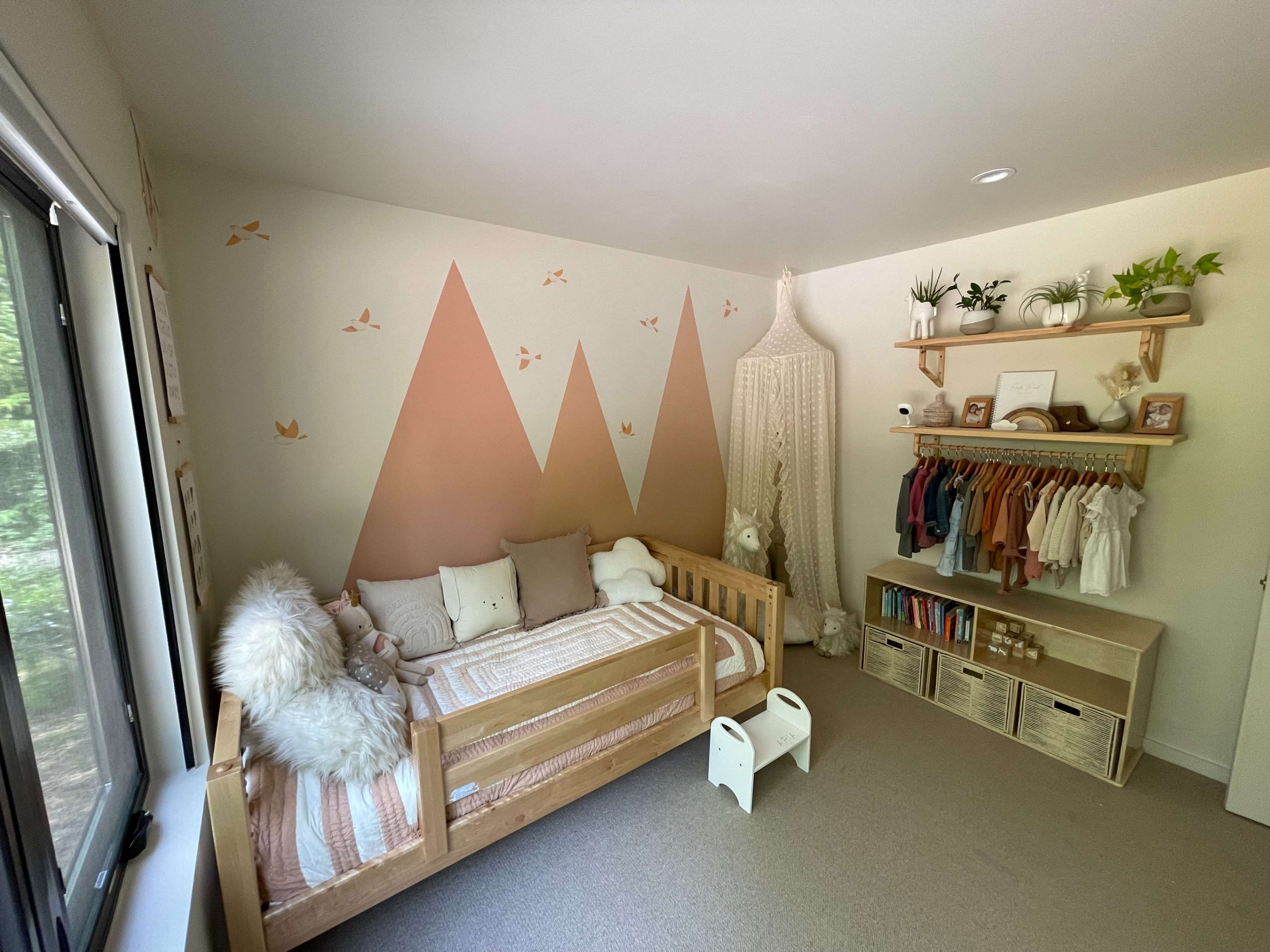 A children's bedroom with a wooden bed, mountain-themed wall decoration, and organized shelves displaying clothing and books.