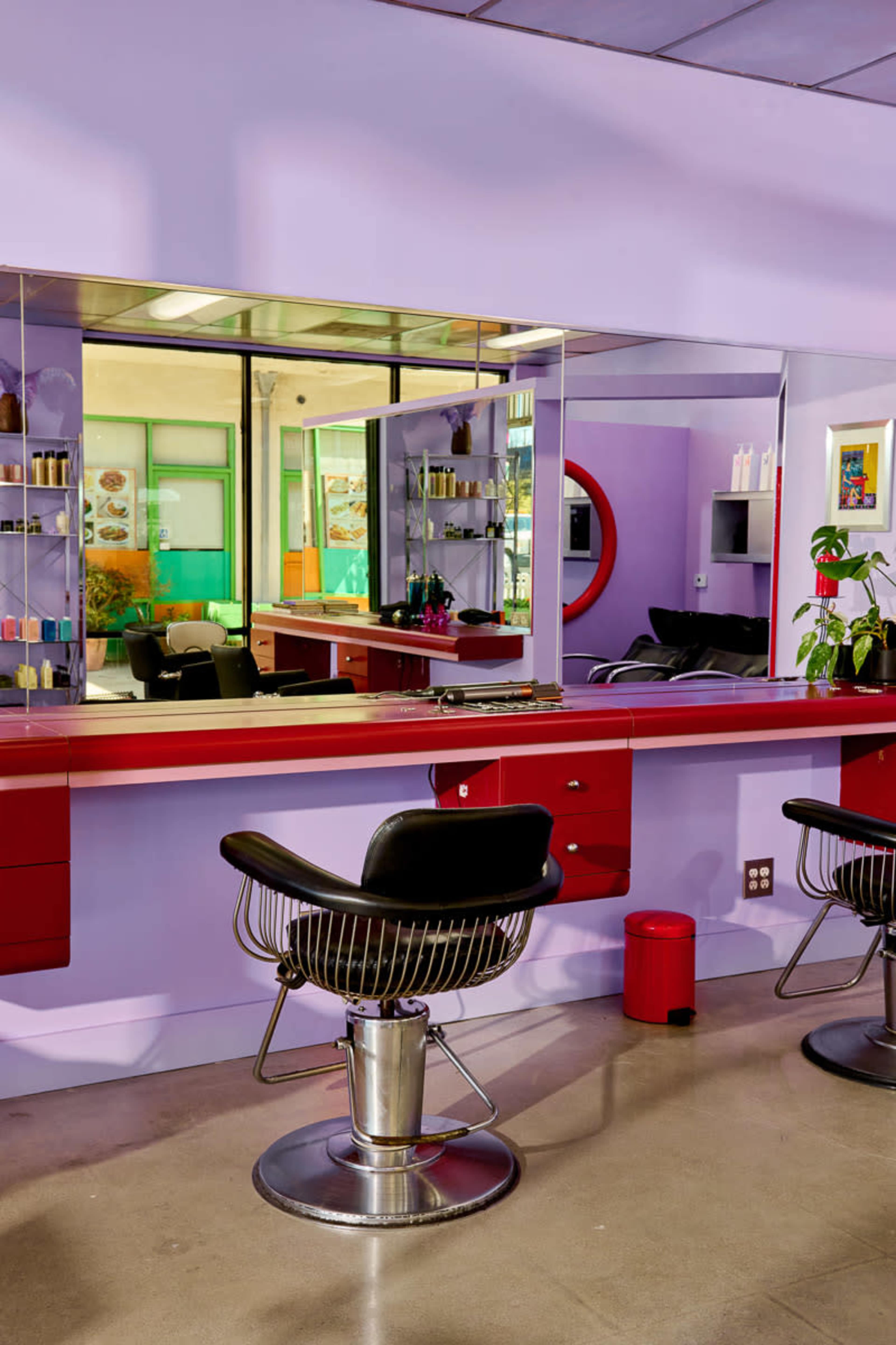 The image shows a brightly lit hair salon featuring two stylist chairs in front of a long mirror, with purple walls and various hair products displayed on shelves.