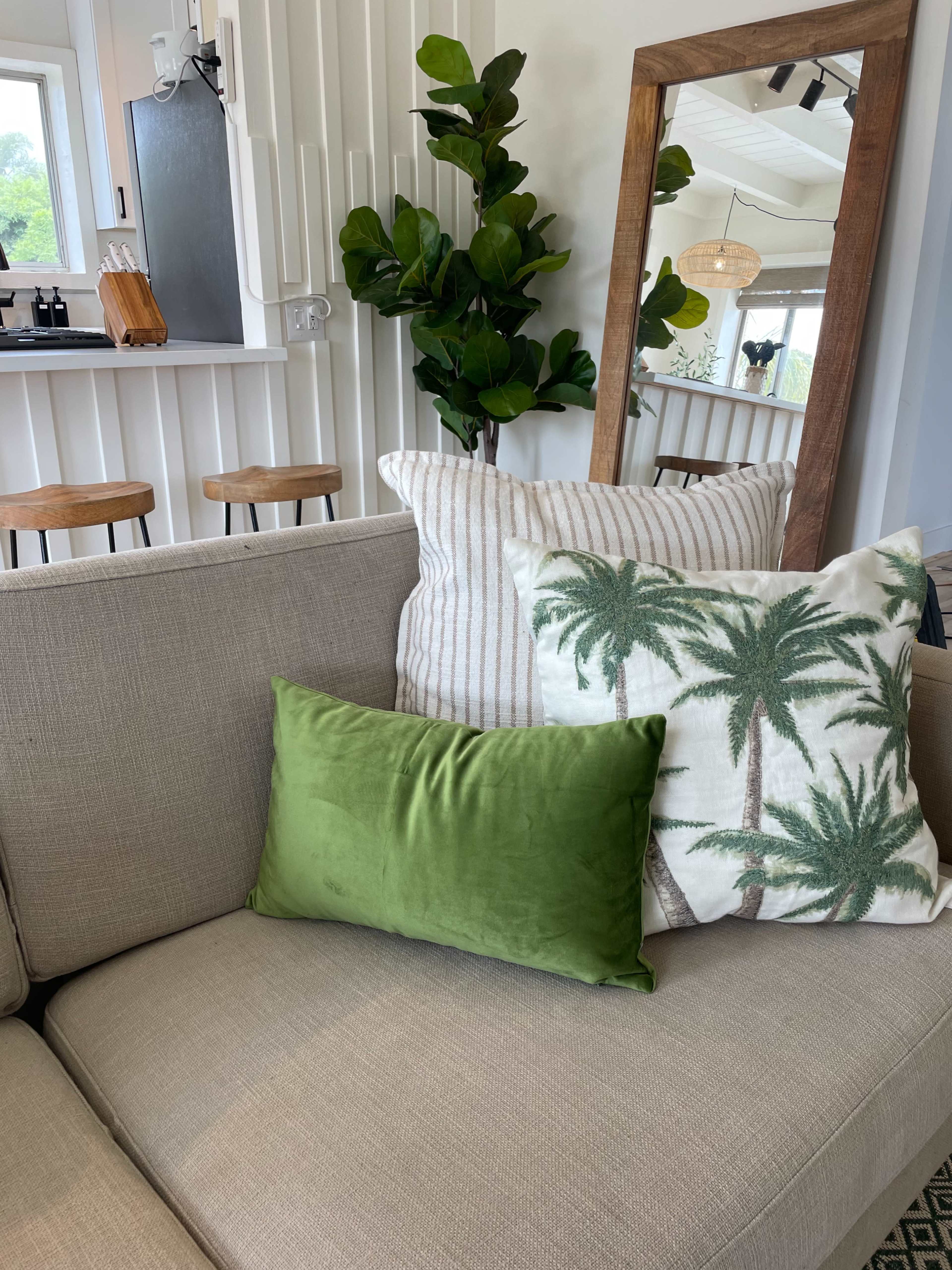 A couch adorned with various decorative pillows, including a green velvet pillow and a palm tree patterned cushion, in a bright, modern interior featuring a large mirror and bar stools in the background.