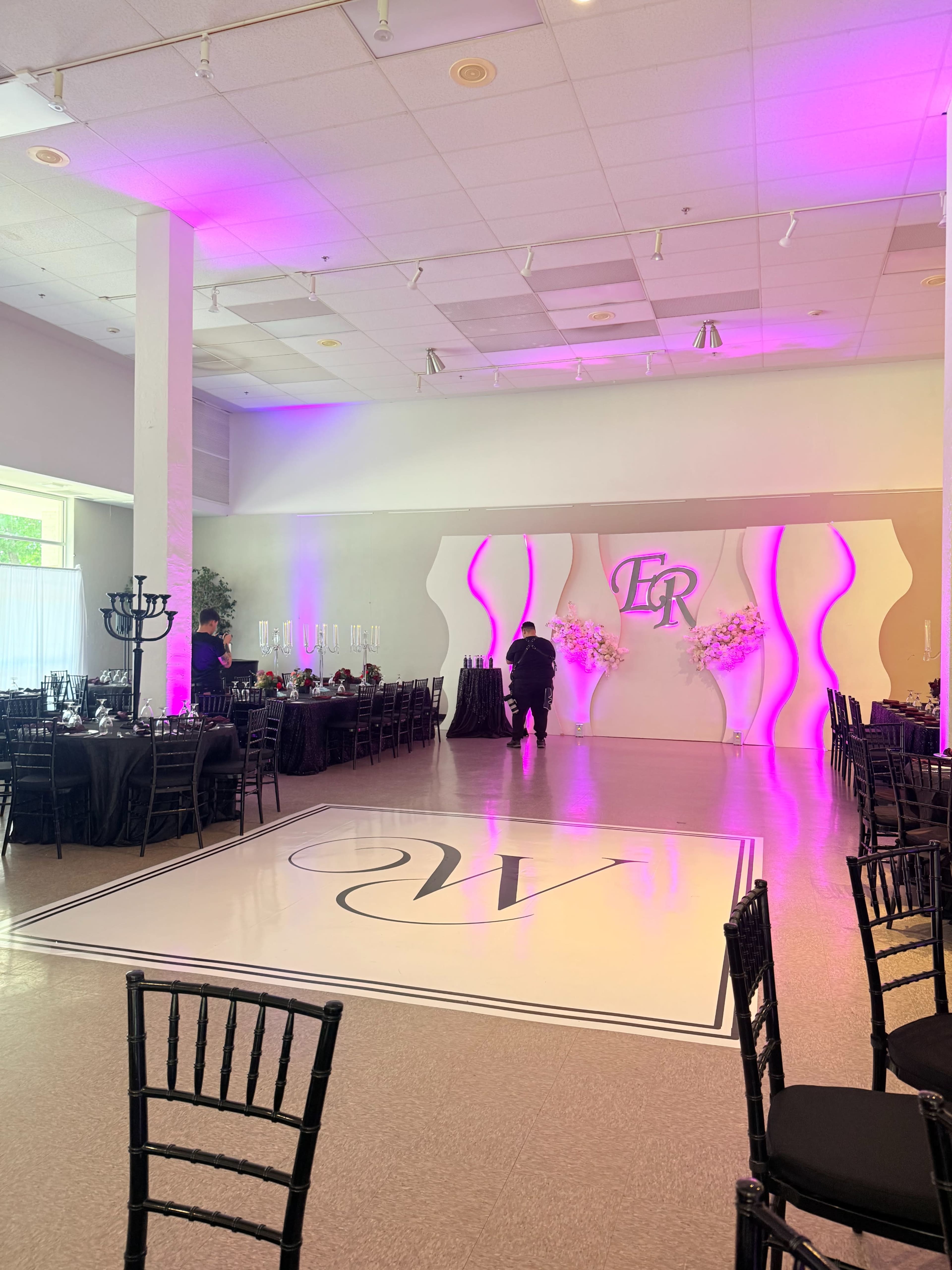 The image shows a banquet hall set up for an event, featuring elegantly arranged tables and a central dance floor with decorative lighting and a backdrop.