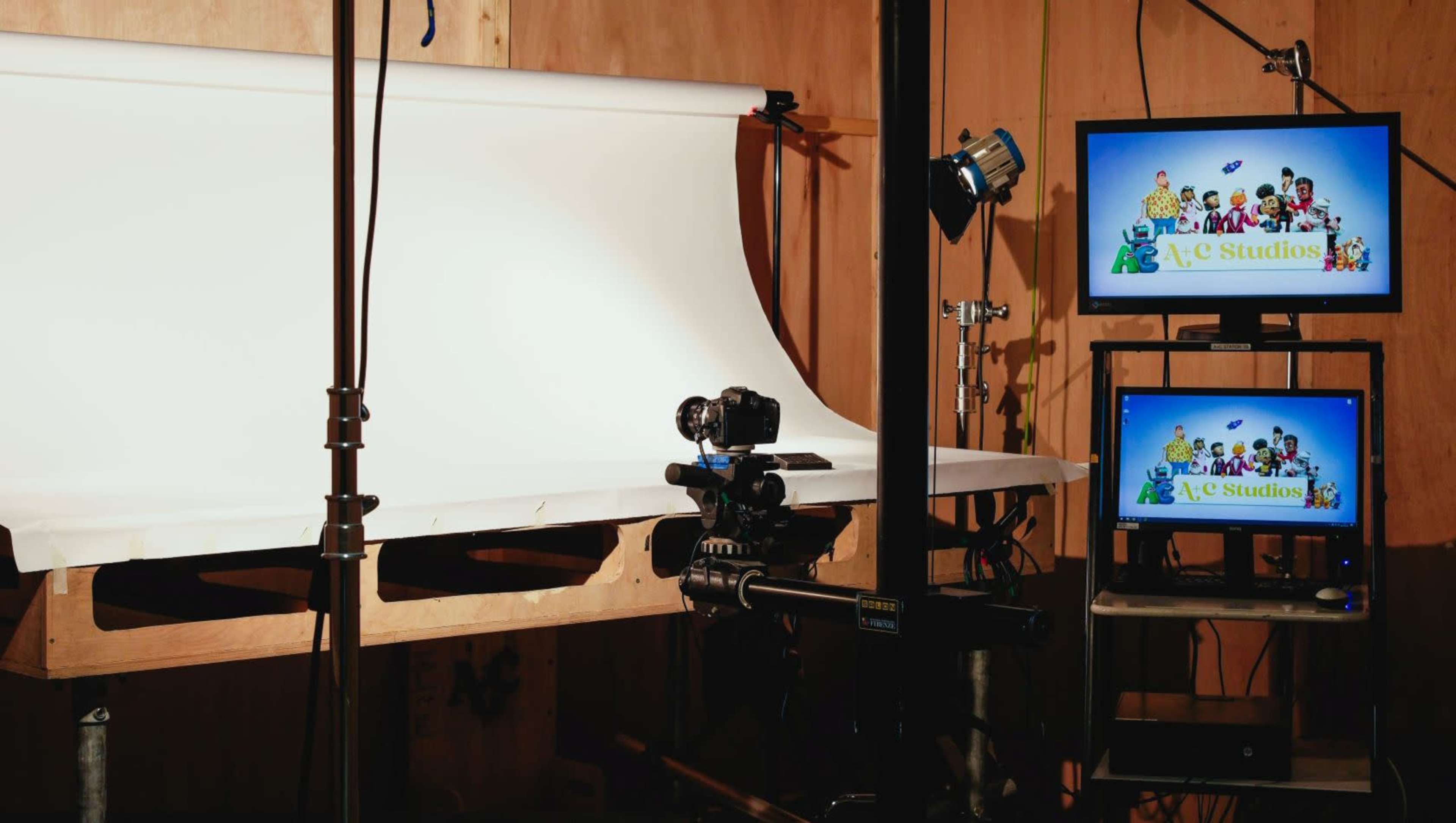 A photography studio setup features a white backdrop, a camera on a tripod, and two monitors displaying colorful characters and the studio name.