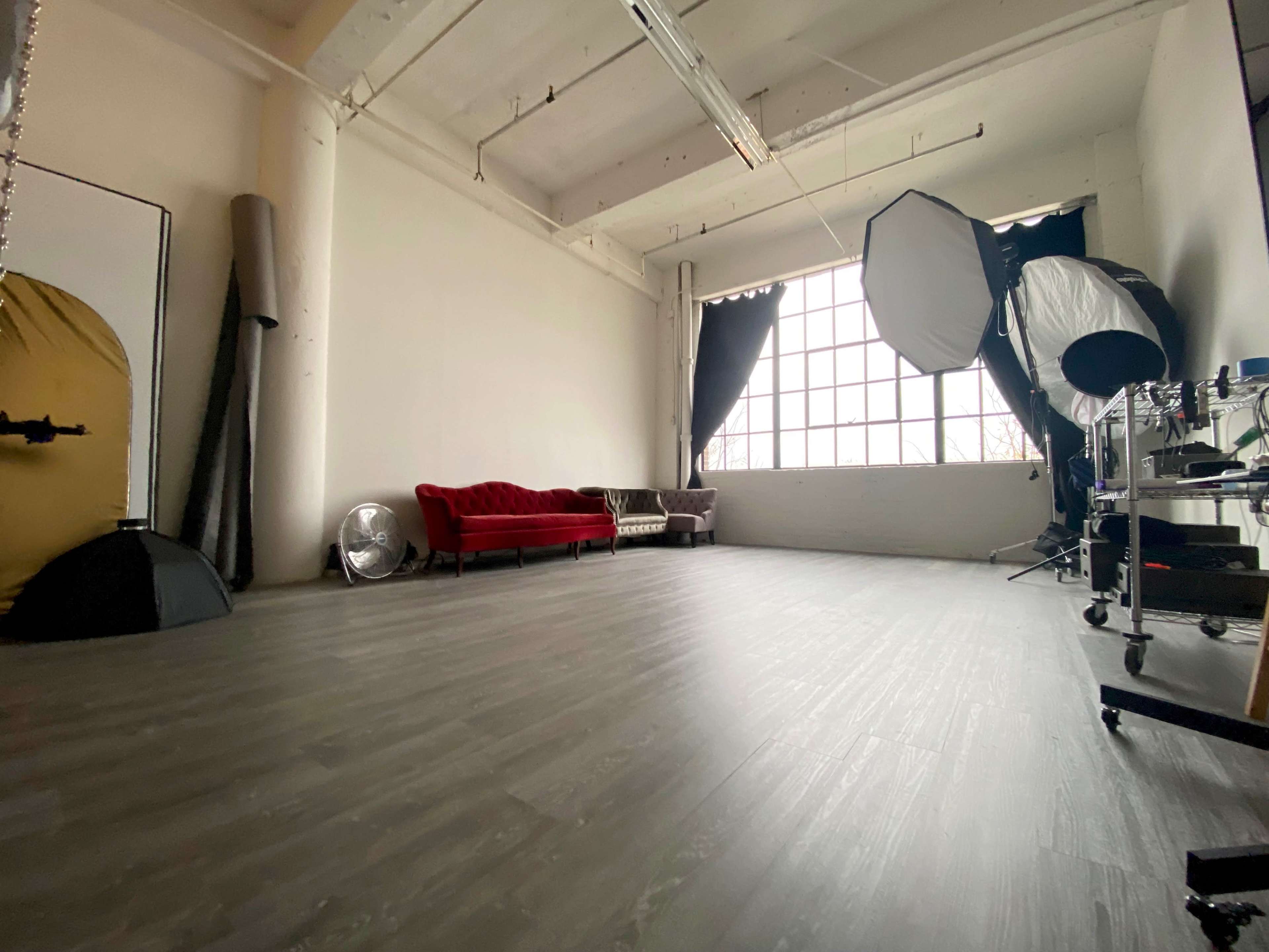 The image features a spacious photography studio with a red velvet sofa, a large window, and various lighting equipment.