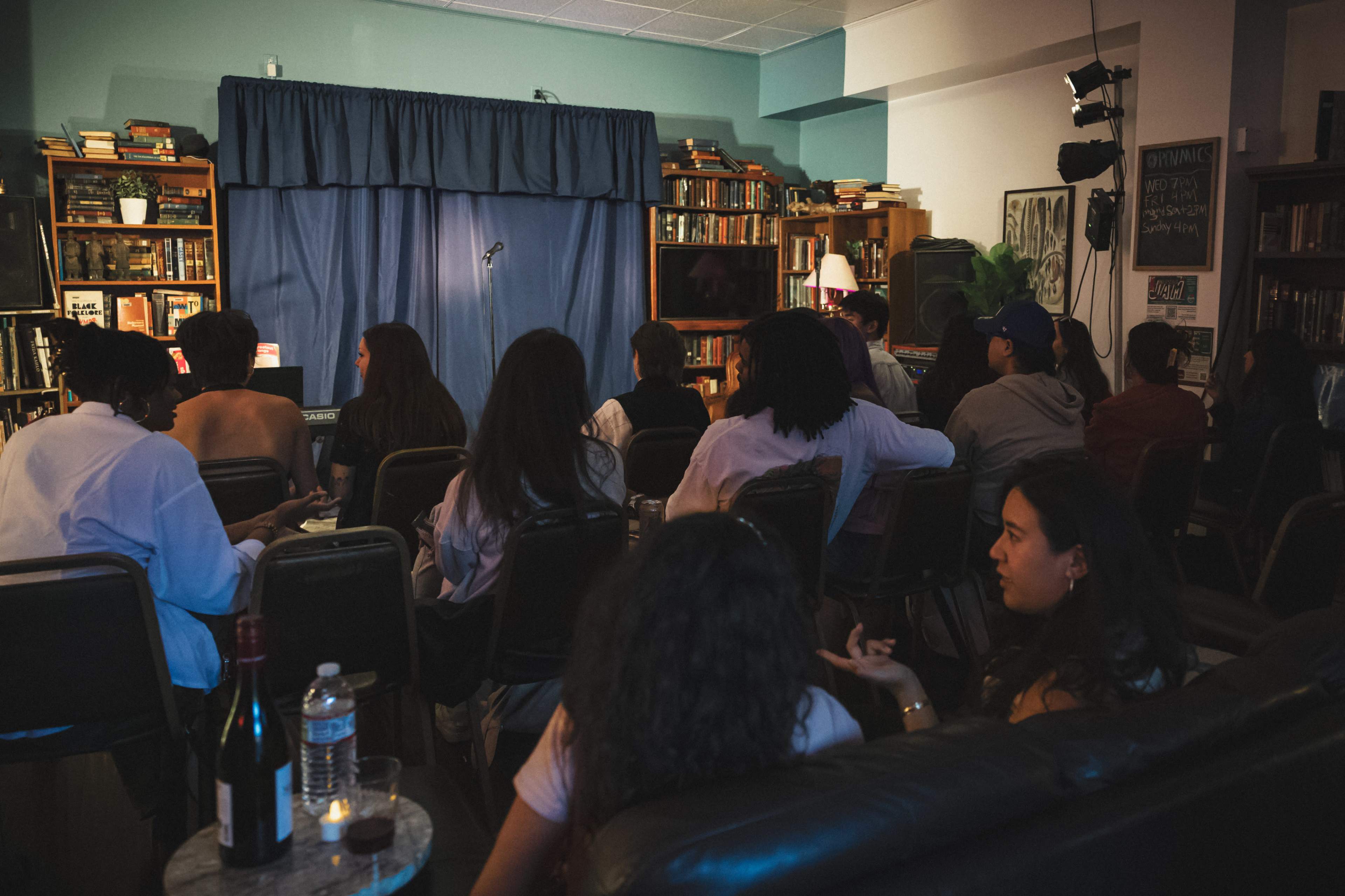 A group of people sits in a small audience facing a stage with a microphone, surrounded by bookshelves and dim lighting.