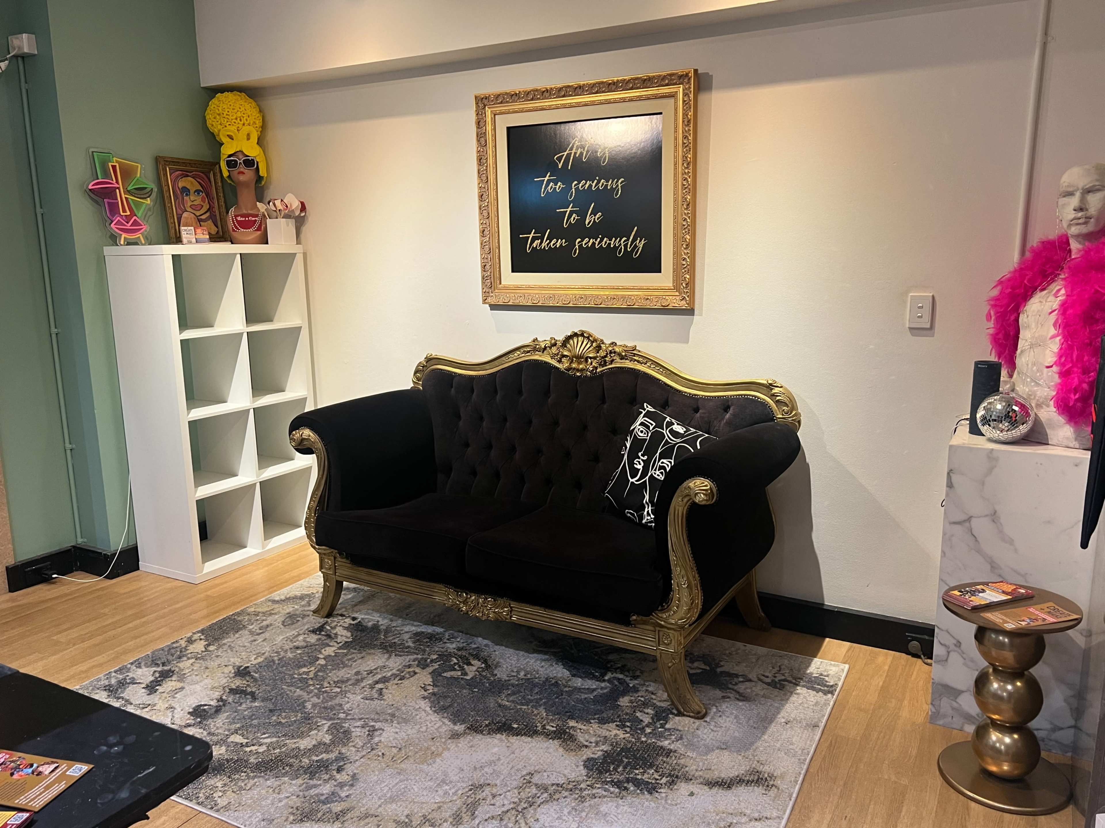 The image shows an ornate black velvet sofa with gold accents, placed on a patterned rug in a brightly lit room with colorful wall art and decorative accessories.