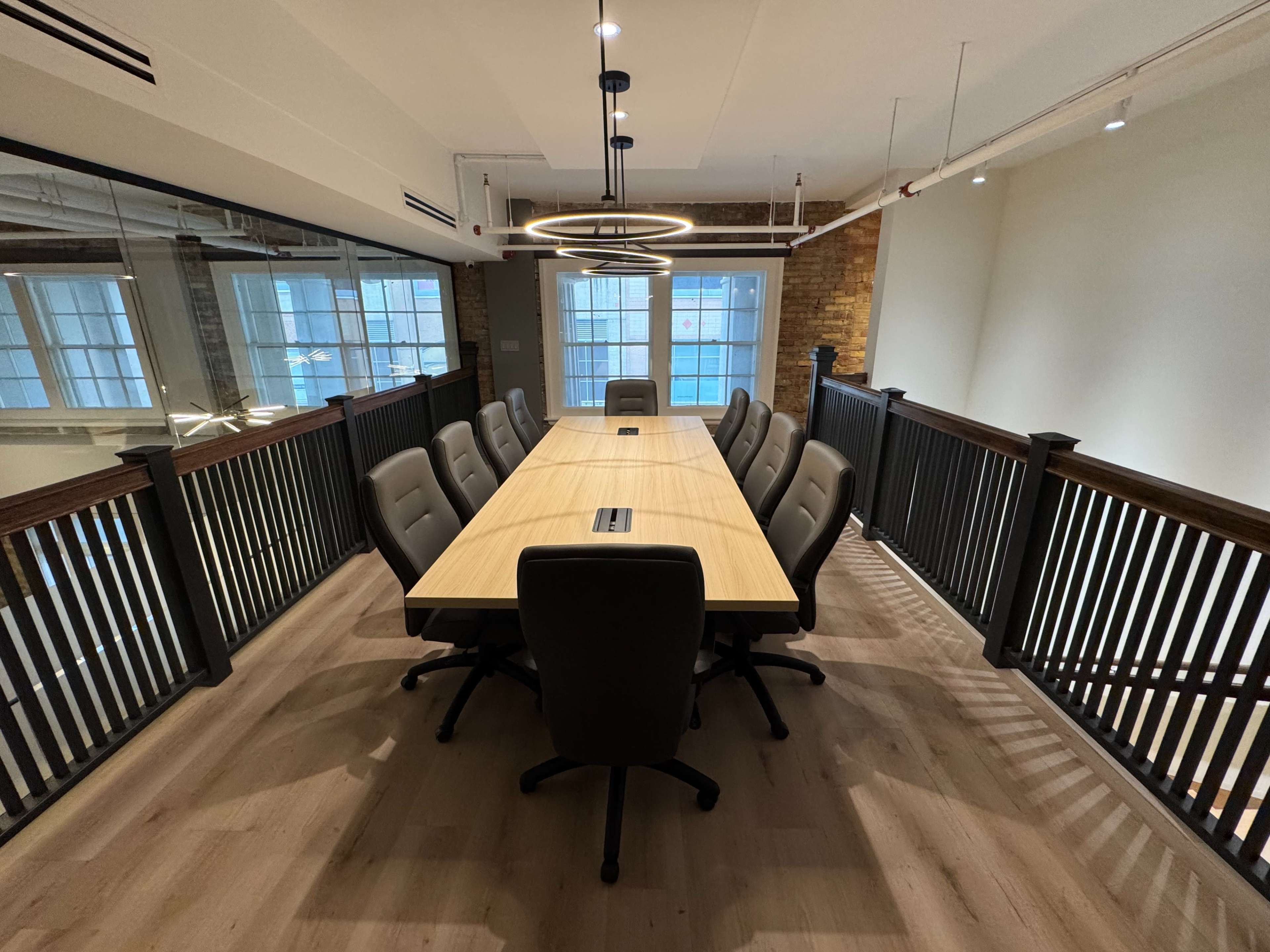 A large conference table with ergonomic chairs is positioned in a well-lit meeting room featuring large windows and exposed brick walls.
