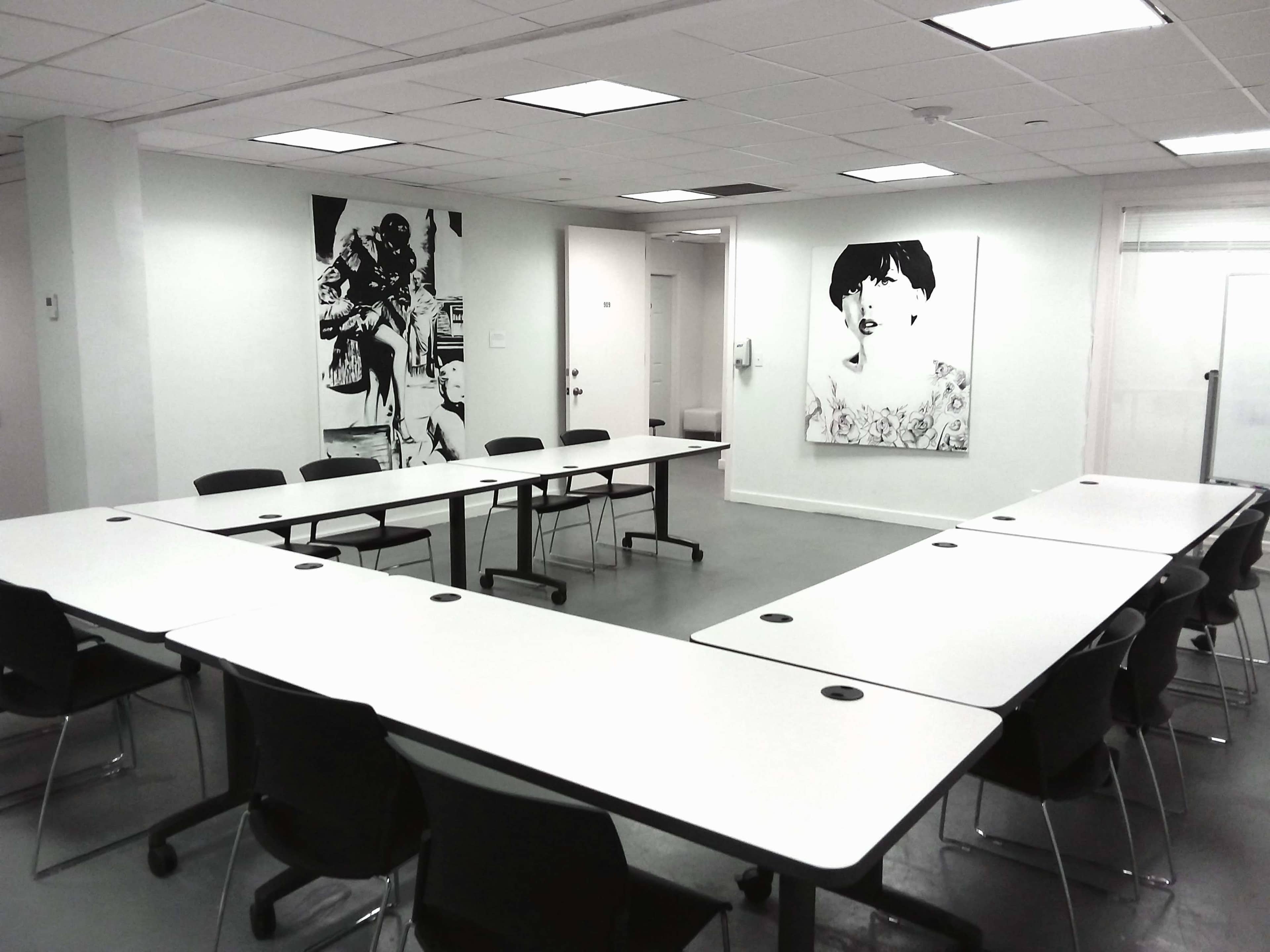 A conference room features a U-shaped arrangement of white tables and black chairs, with large framed artworks on the walls.