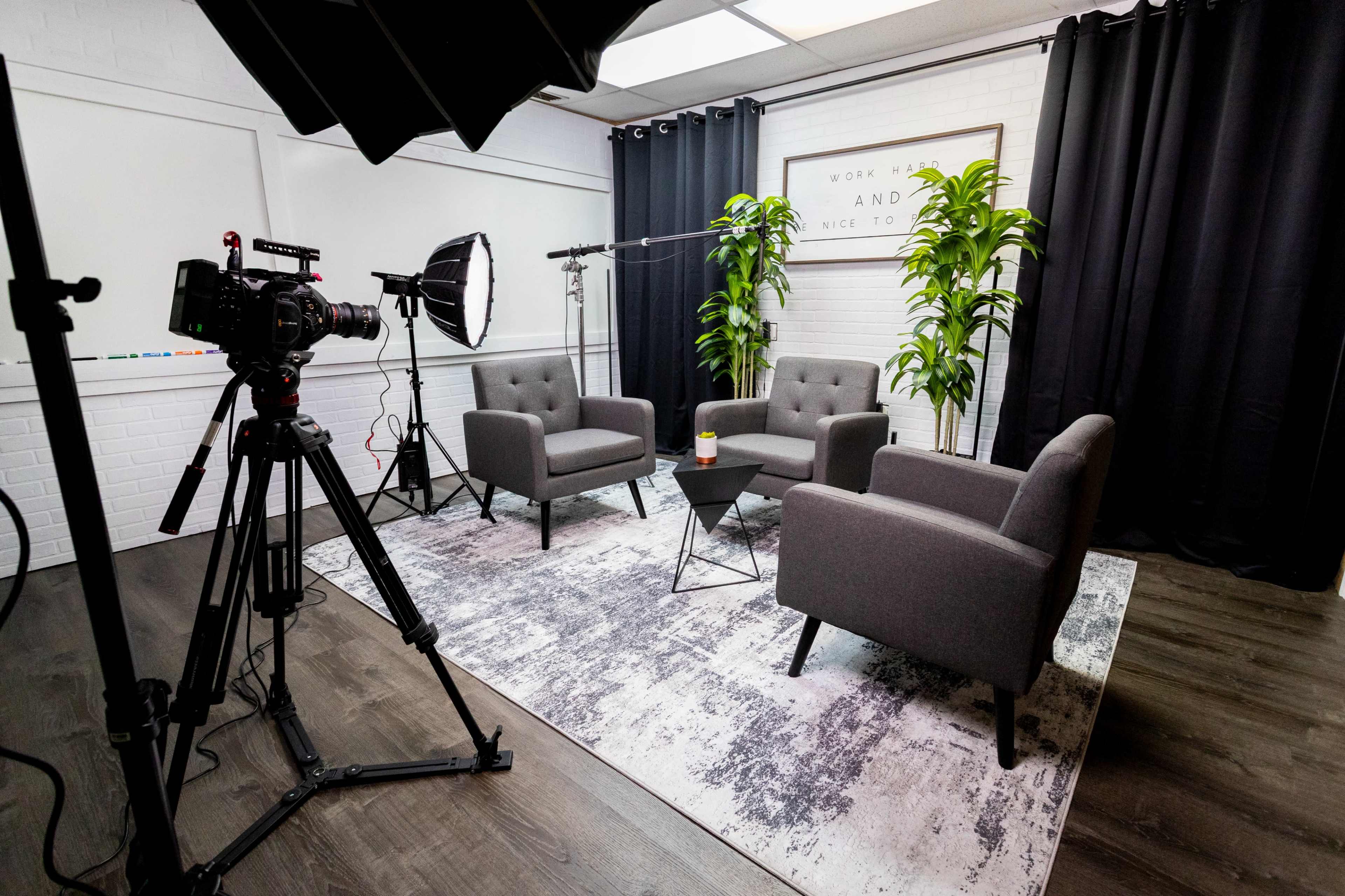 A well-lit studio setting features two gray armchairs, a small black table, and potted plants against a backdrop of dark curtains and a white-brick wall.