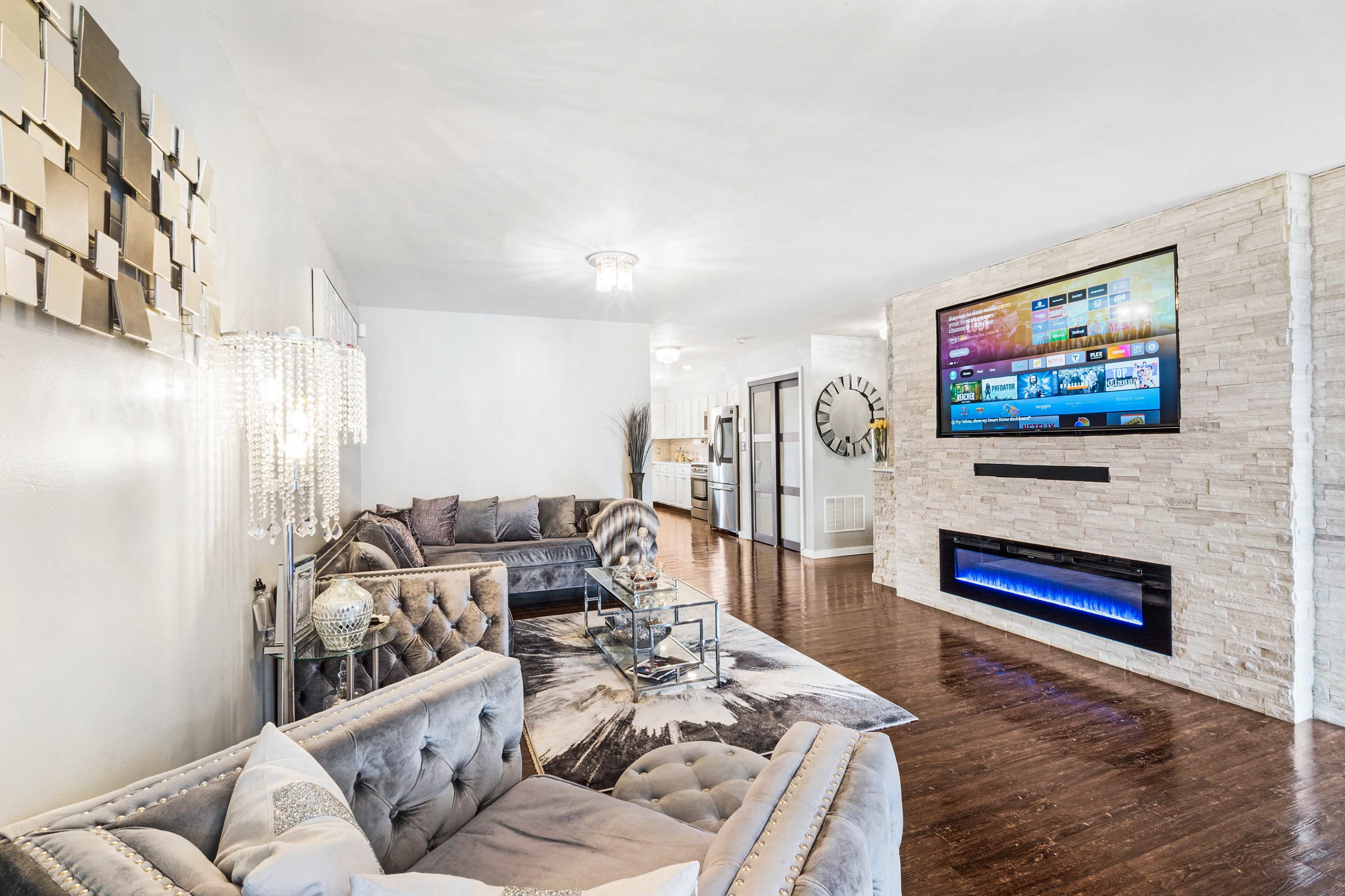 The living room features modern furniture with a large TV mounted on a stone wall and a fireplace below it.