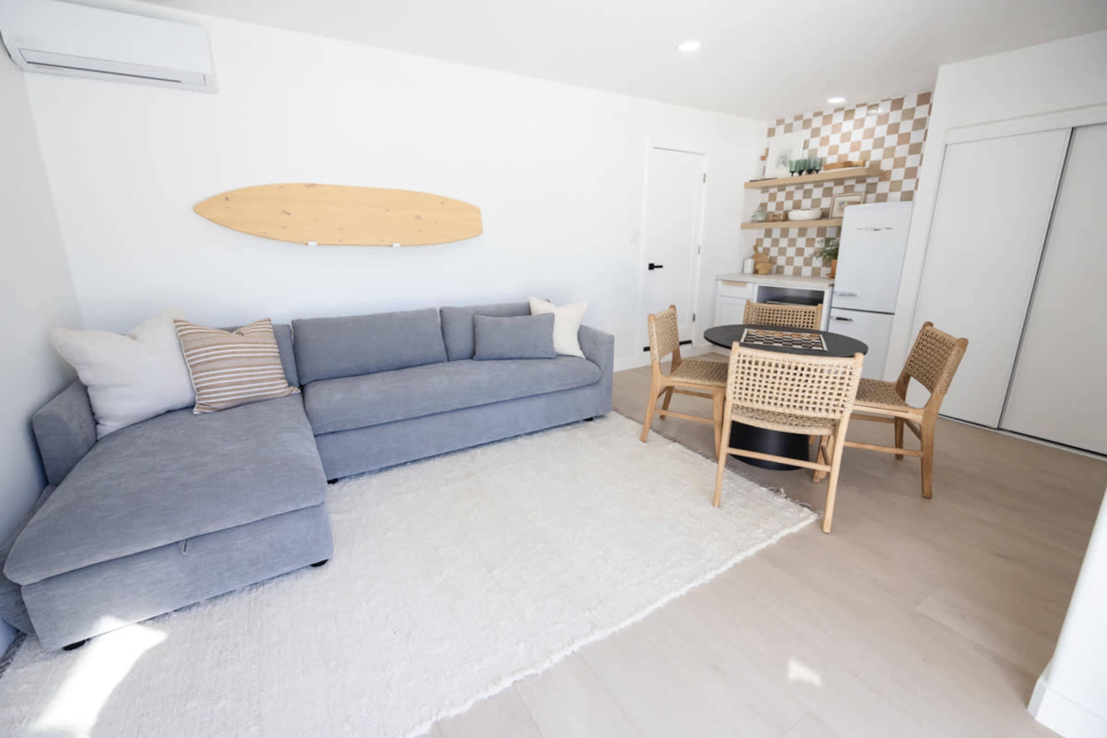 A bright, minimalist living space featuring a gray sectional sofa, a round dining table with chairs, and a kitchenette with a patterned backsplash.