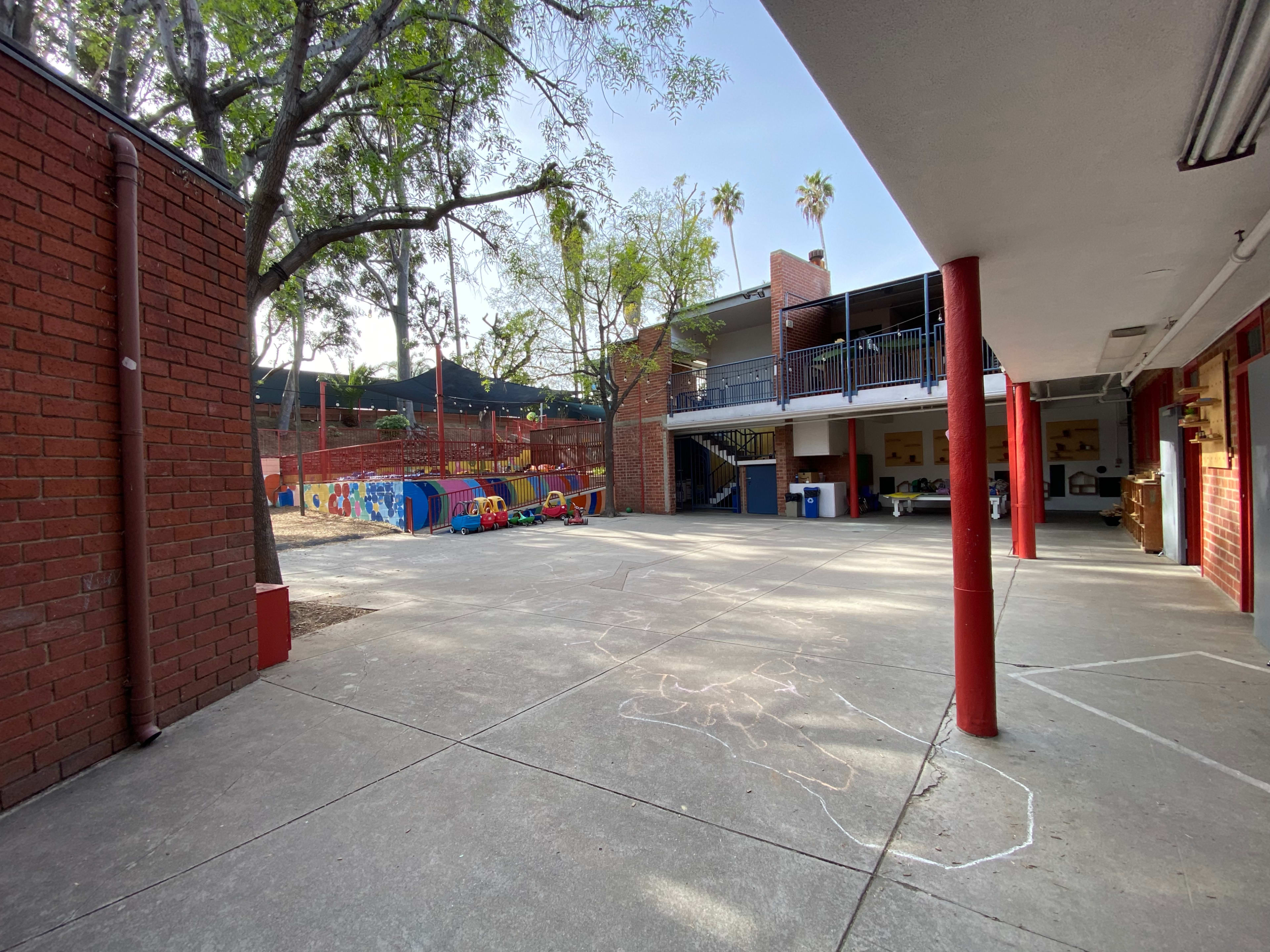 A spacious school courtyard with red brick walls, a playground area in the background, and trees providing some shade.