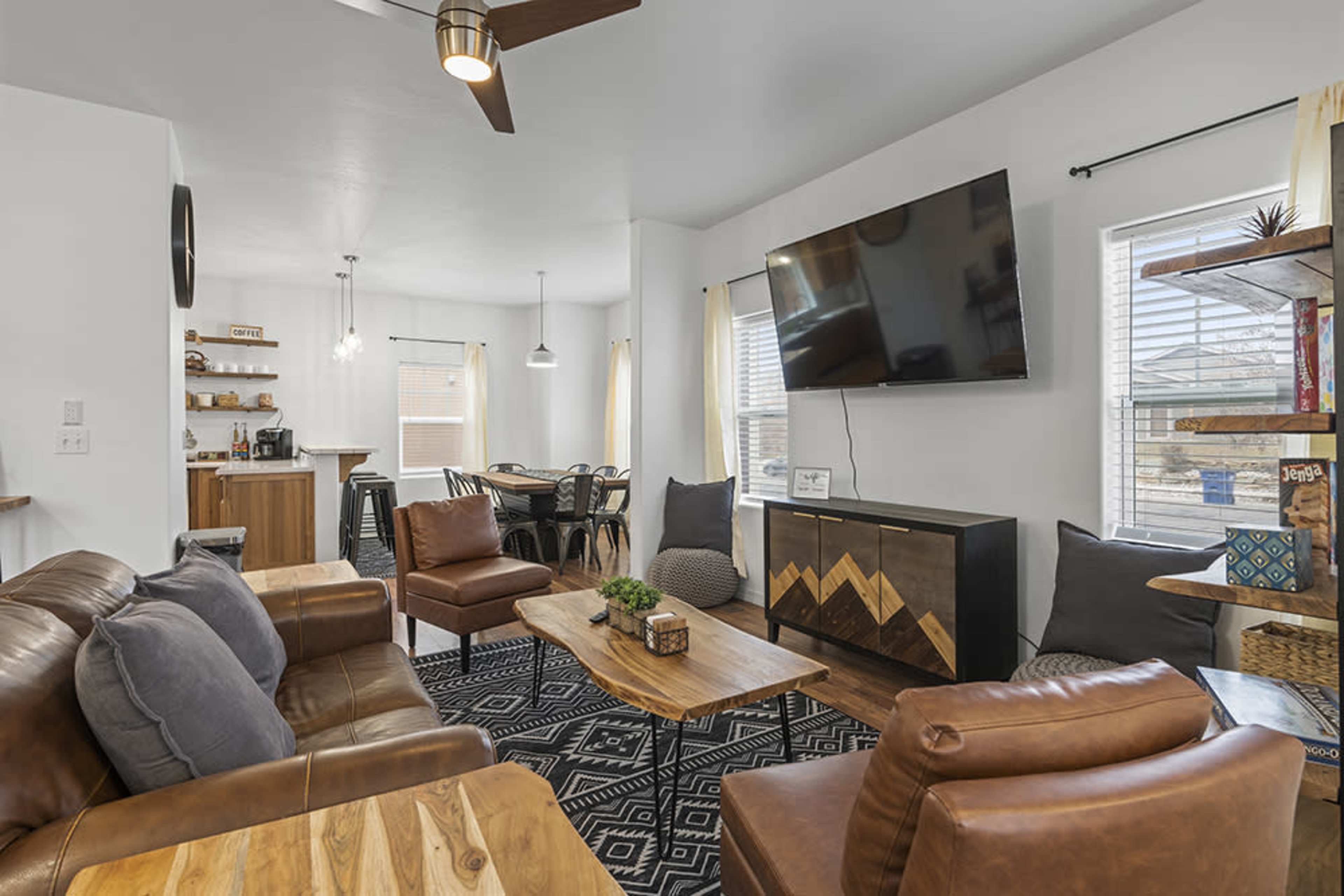 A modern living room featuring a sectional sofa, two armchairs, a coffee table, and a large wall-mounted television, with a dining area visible in the background.