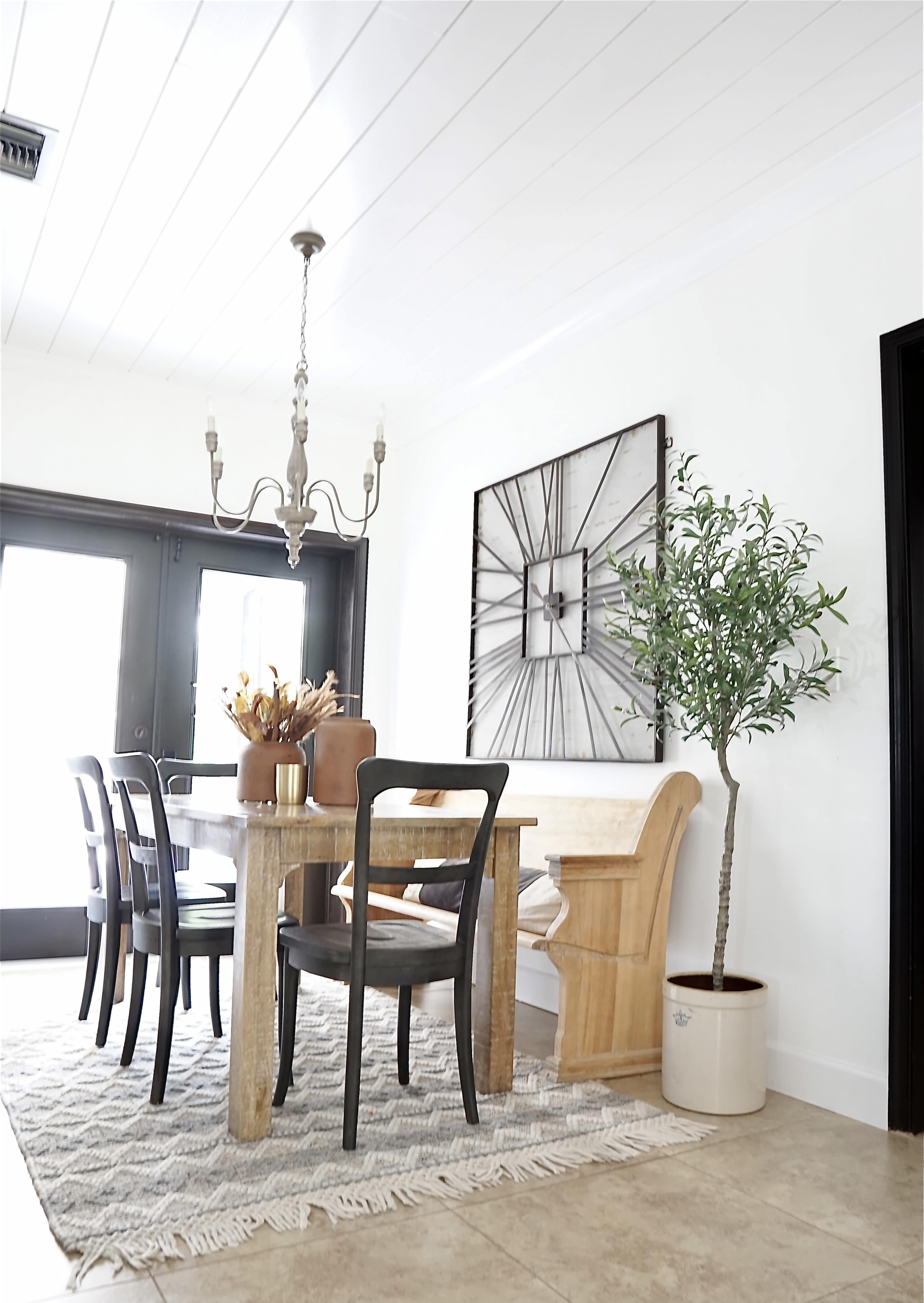 A dining area features a wooden table surrounded by black chairs, with a chandelier overhead and a potted plant in the corner against a light-colored wall.