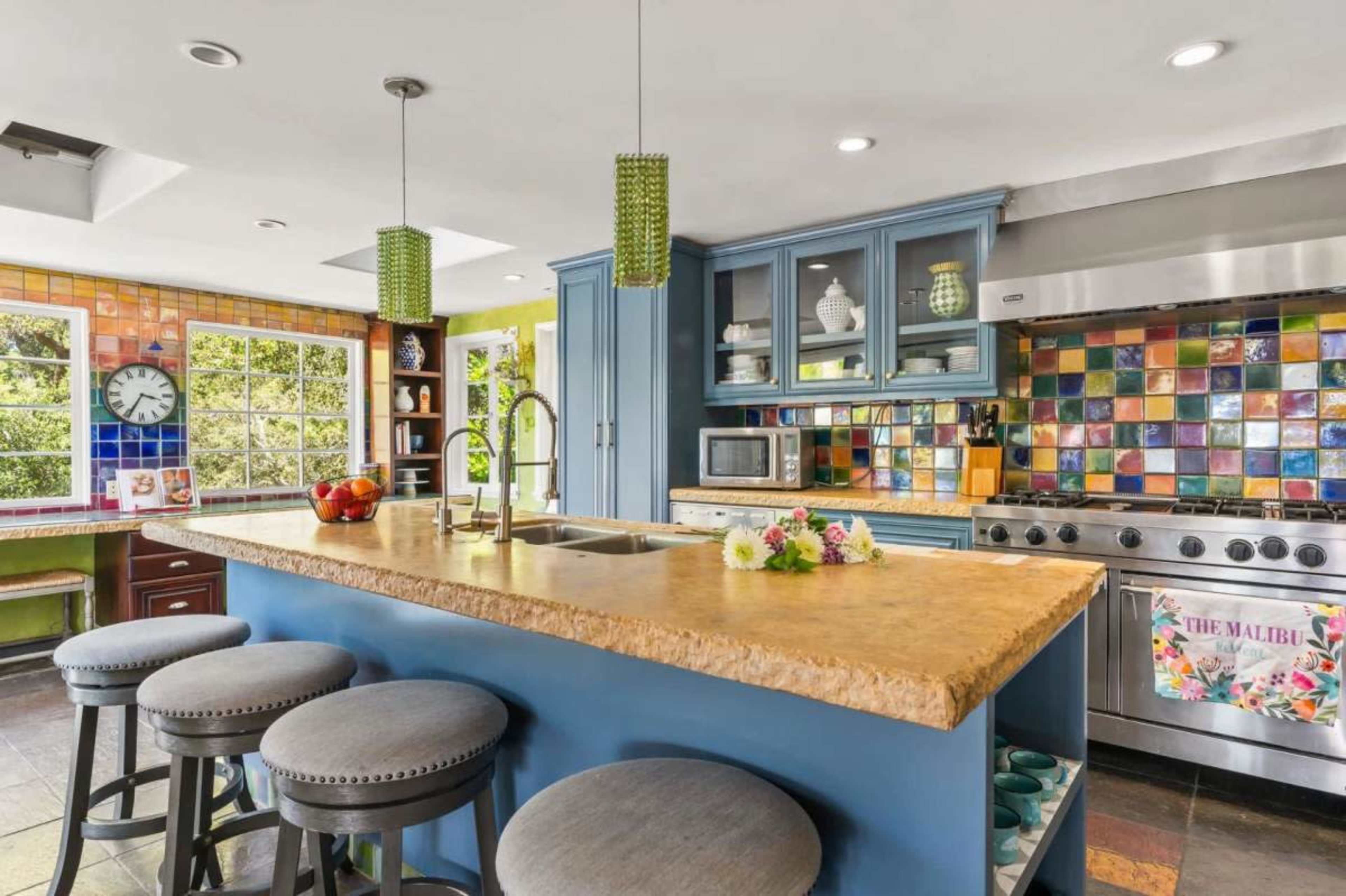 The image shows a modern kitchen with a colorful tiled backsplash, a large island with a stone countertop, and bar stools.