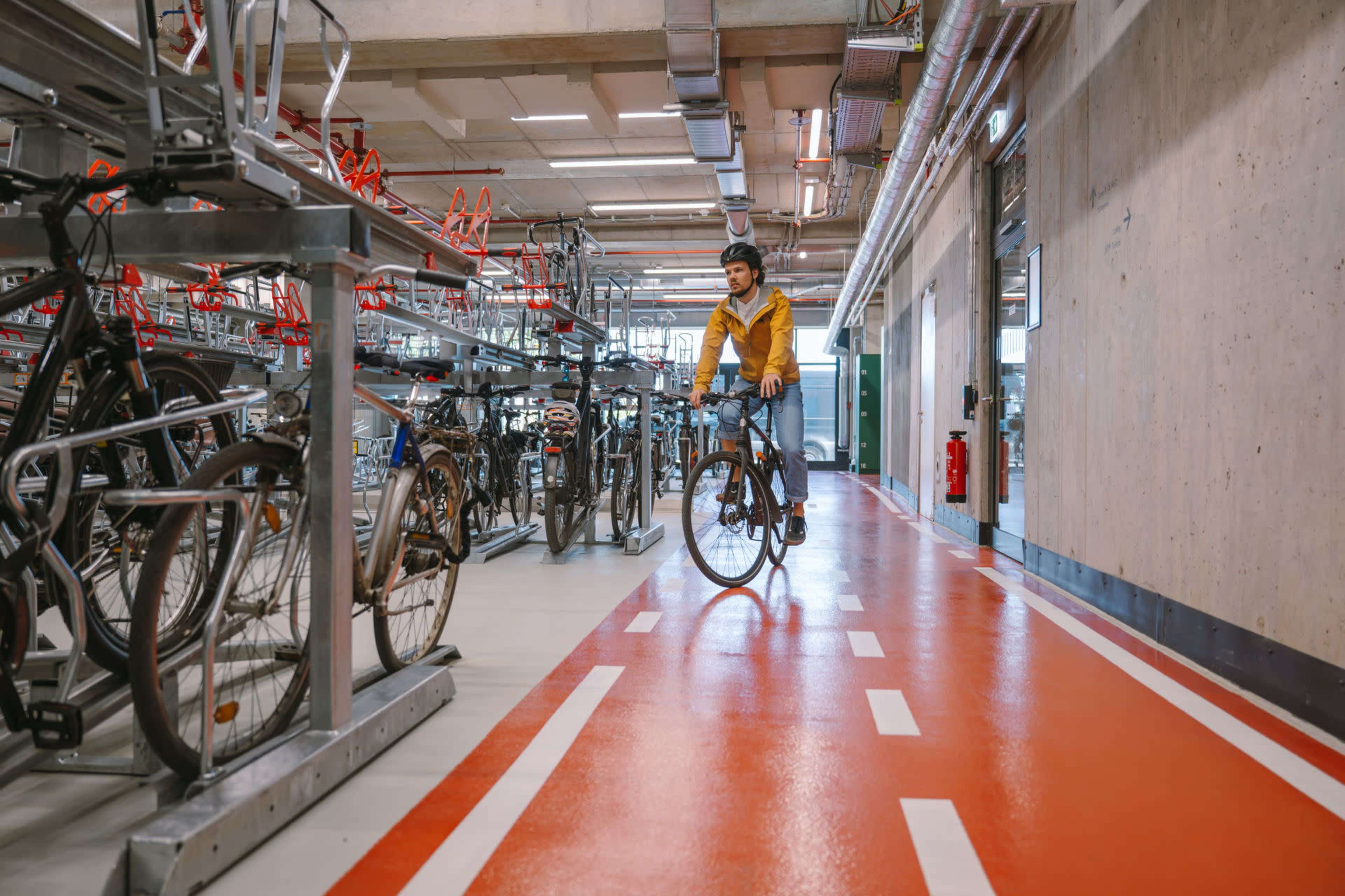 A person wearing a yellow jacket rides a bicycle through a well-organized indoor bike parking area with red and white lanes.