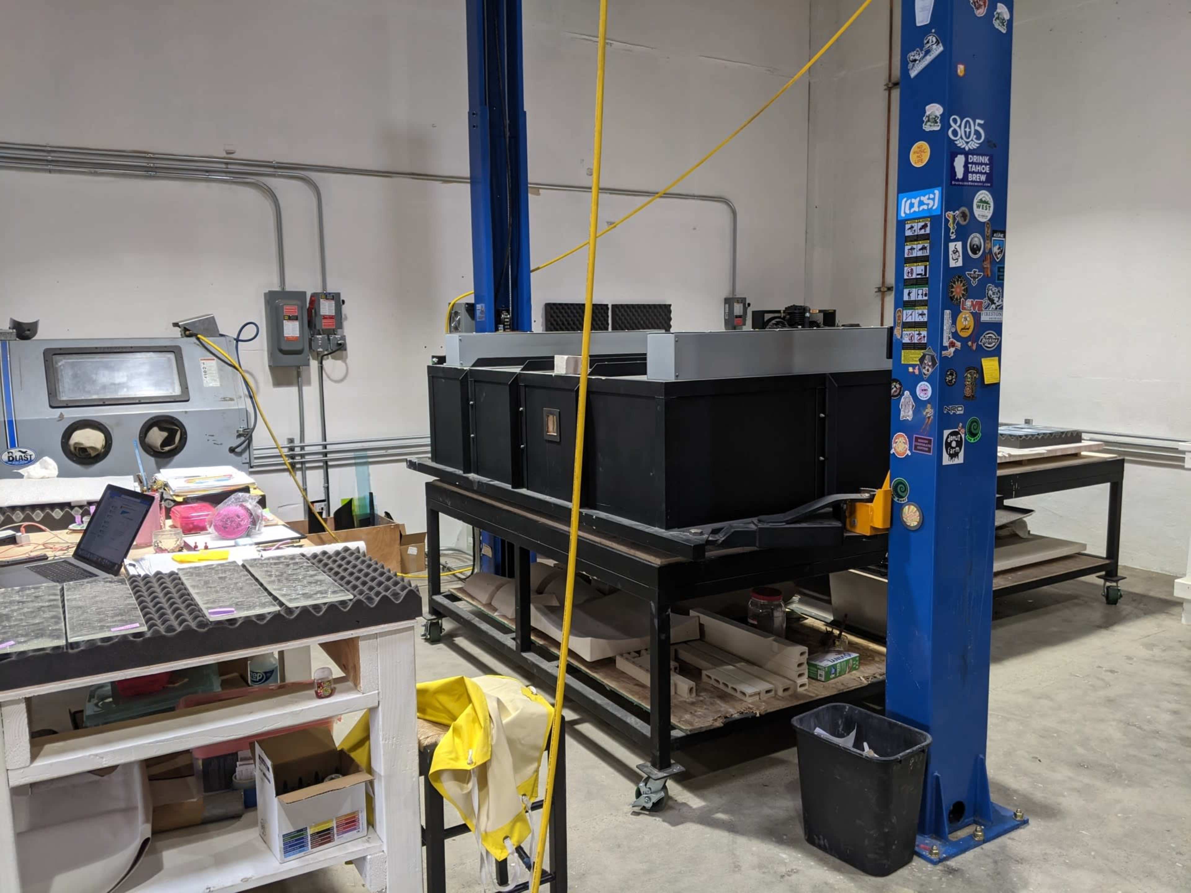 A workshop with a large black machine on a platform, surrounded by various tools and equipment on a workbench and a blue support column.