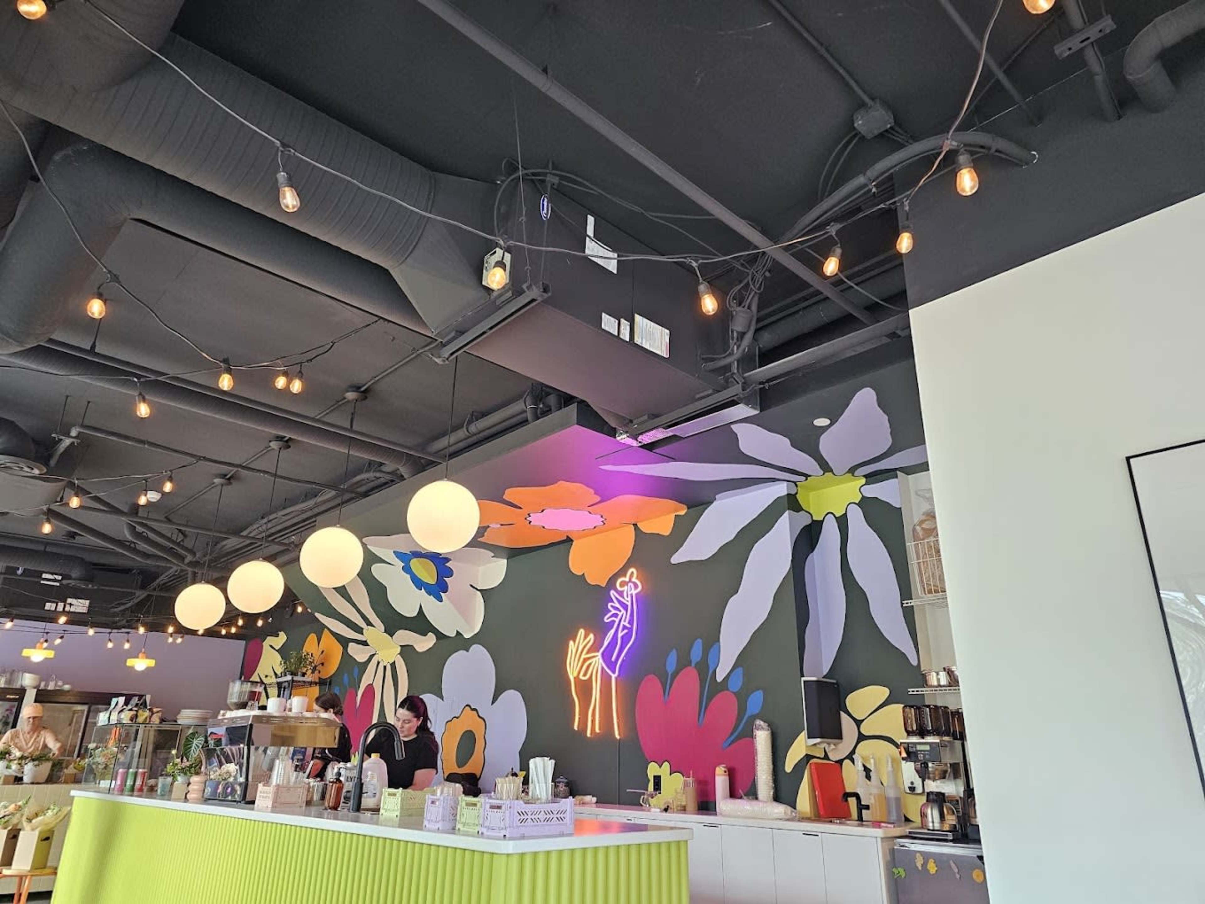 A brightly decorated café features a floral mural and suspended lighting above a service counter.