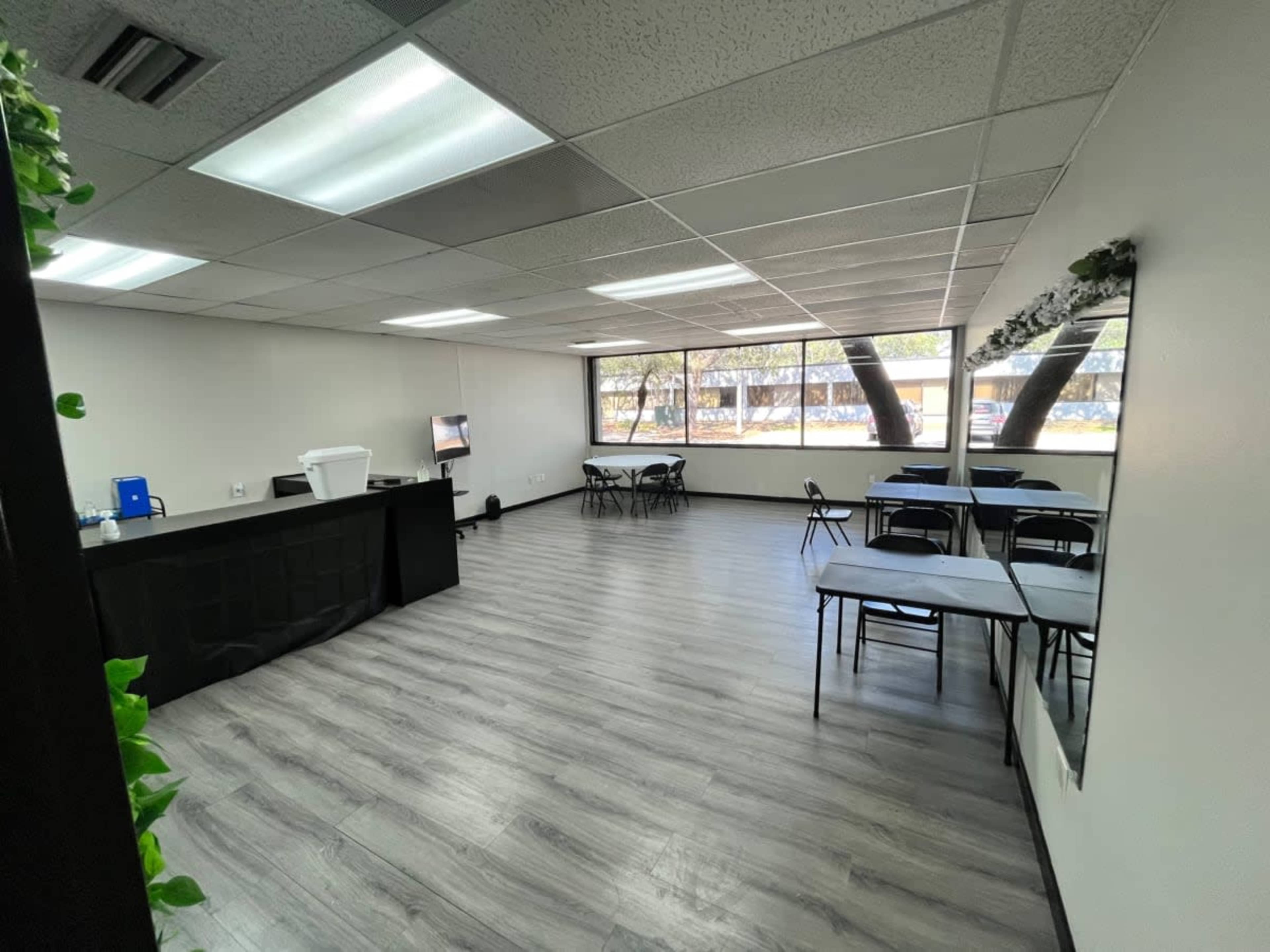 The image shows a spacious, well-lit room with a few tables and chairs arranged, large windows providing natural light, and a black counter against one wall.
