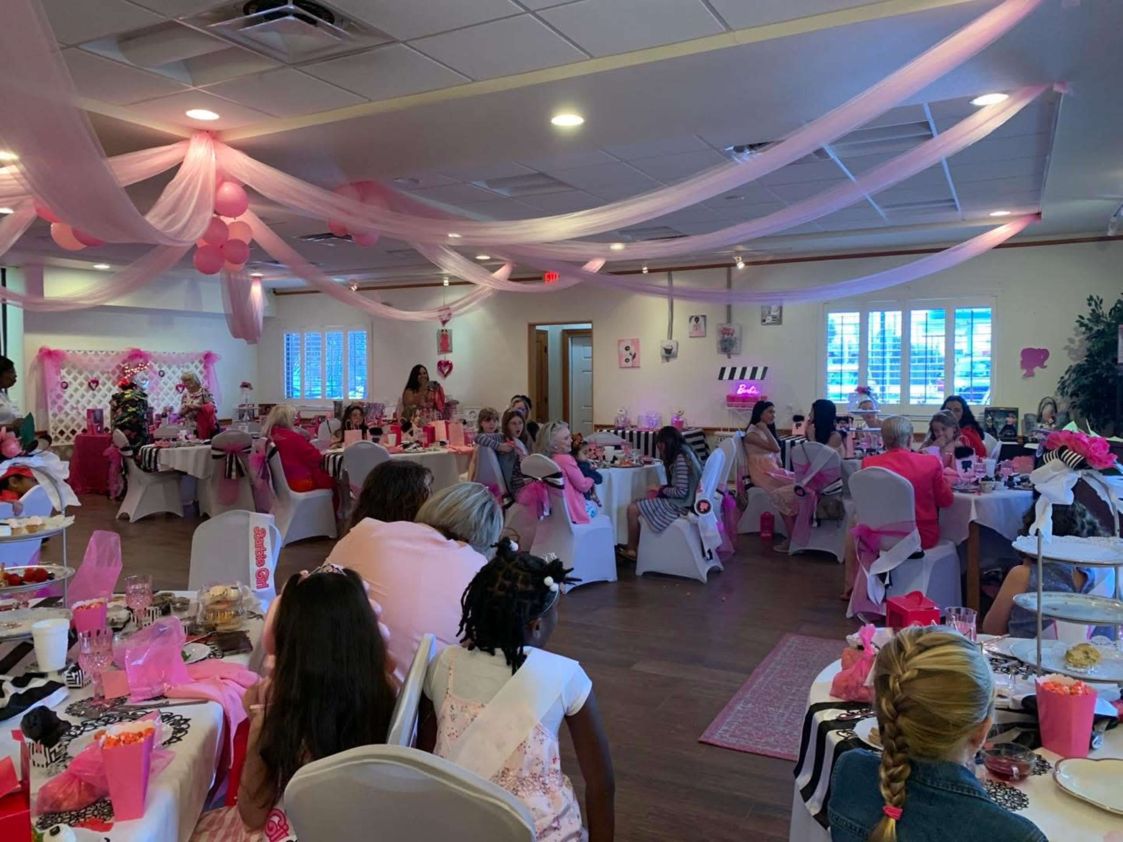 A gathering of children and adults dressed in pink is taking place in a decorated event space with tables set and pink-themed decorations overhead.