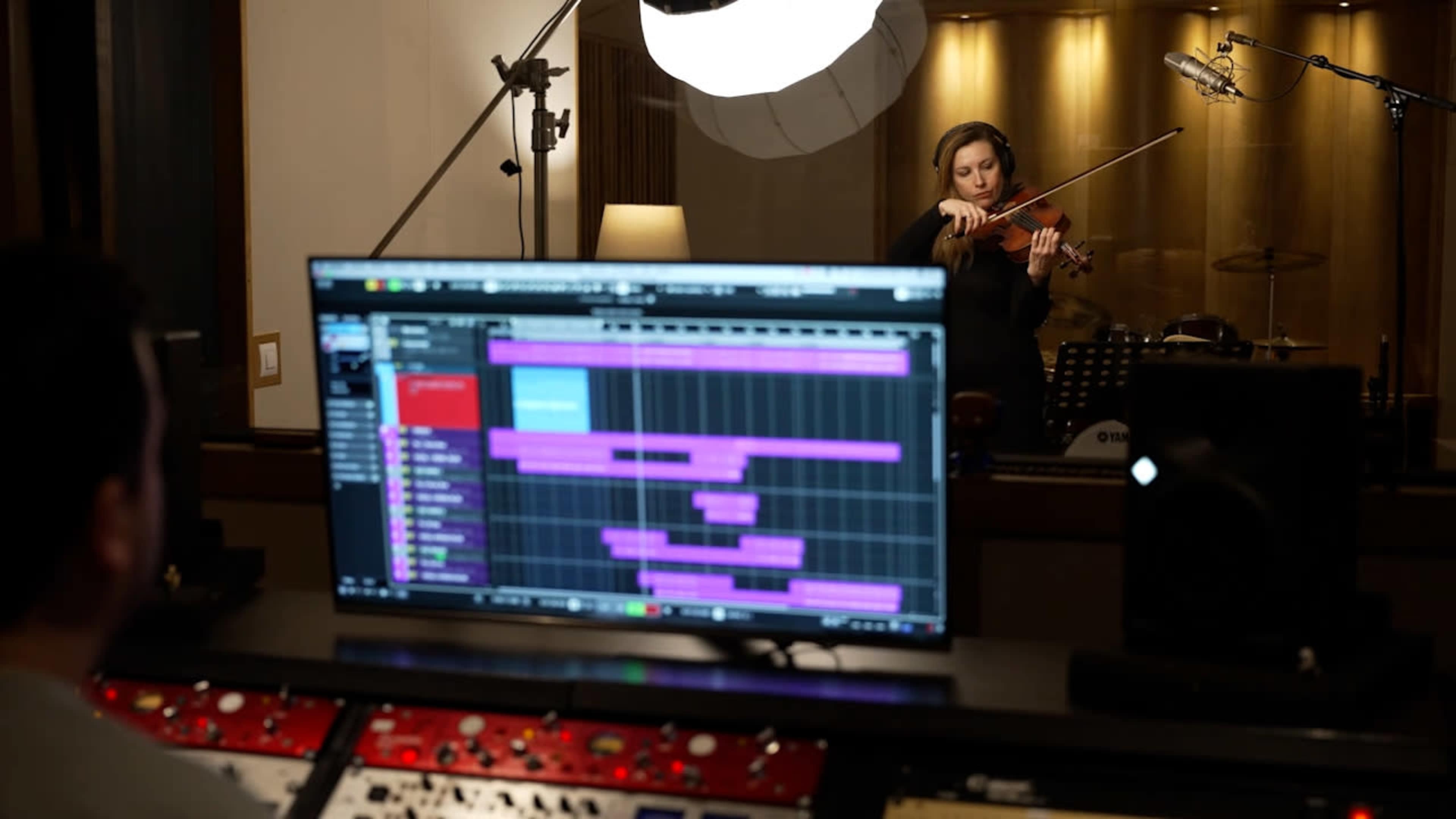 A musician plays the violin in a recording studio while a sound engineer monitors the session from a computer screen displaying audio tracks.