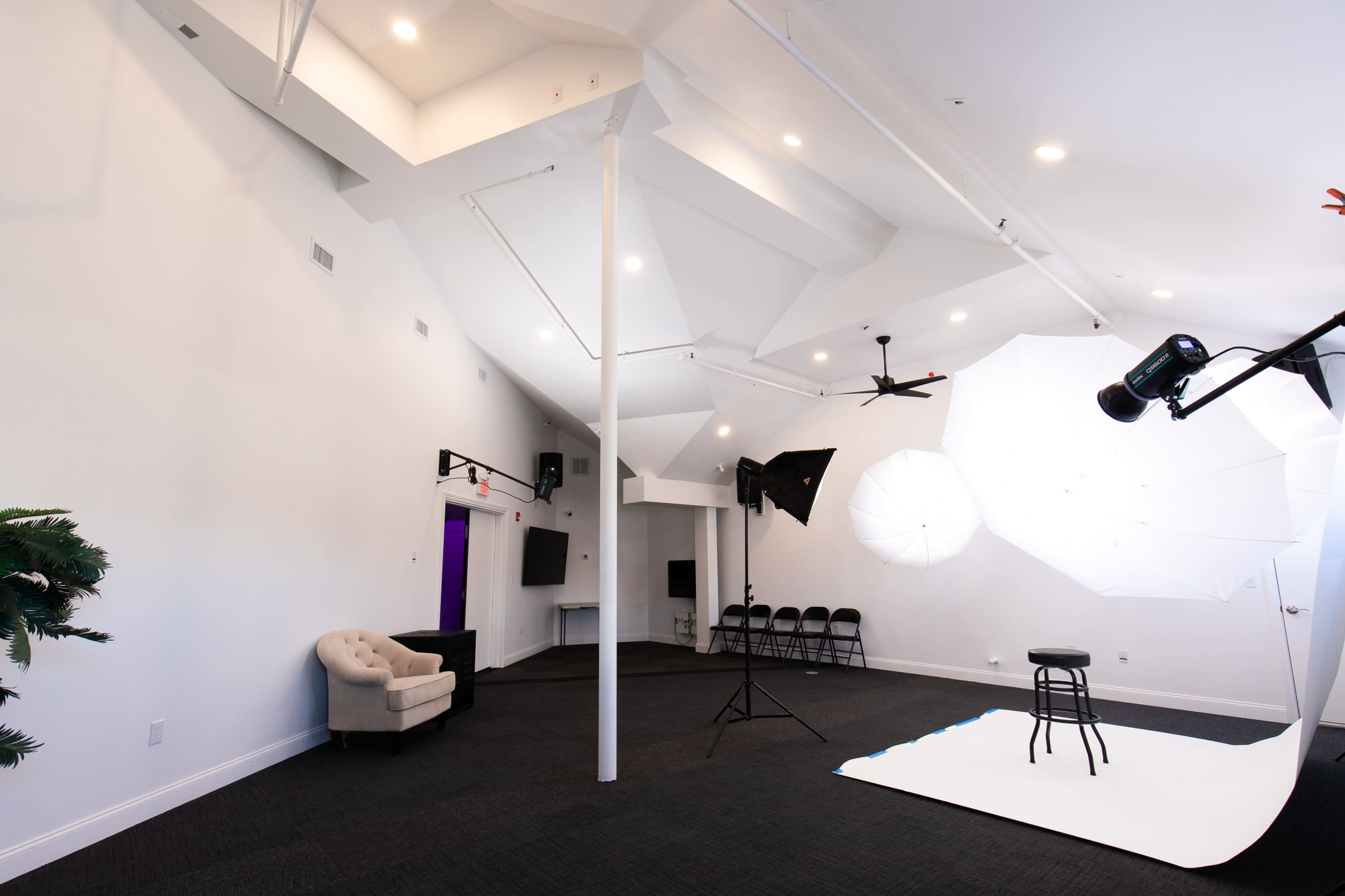 The image shows a spacious photo studio with soft lighting equipment, a stool on a white backdrop, and a few black chairs lined up against the wall.