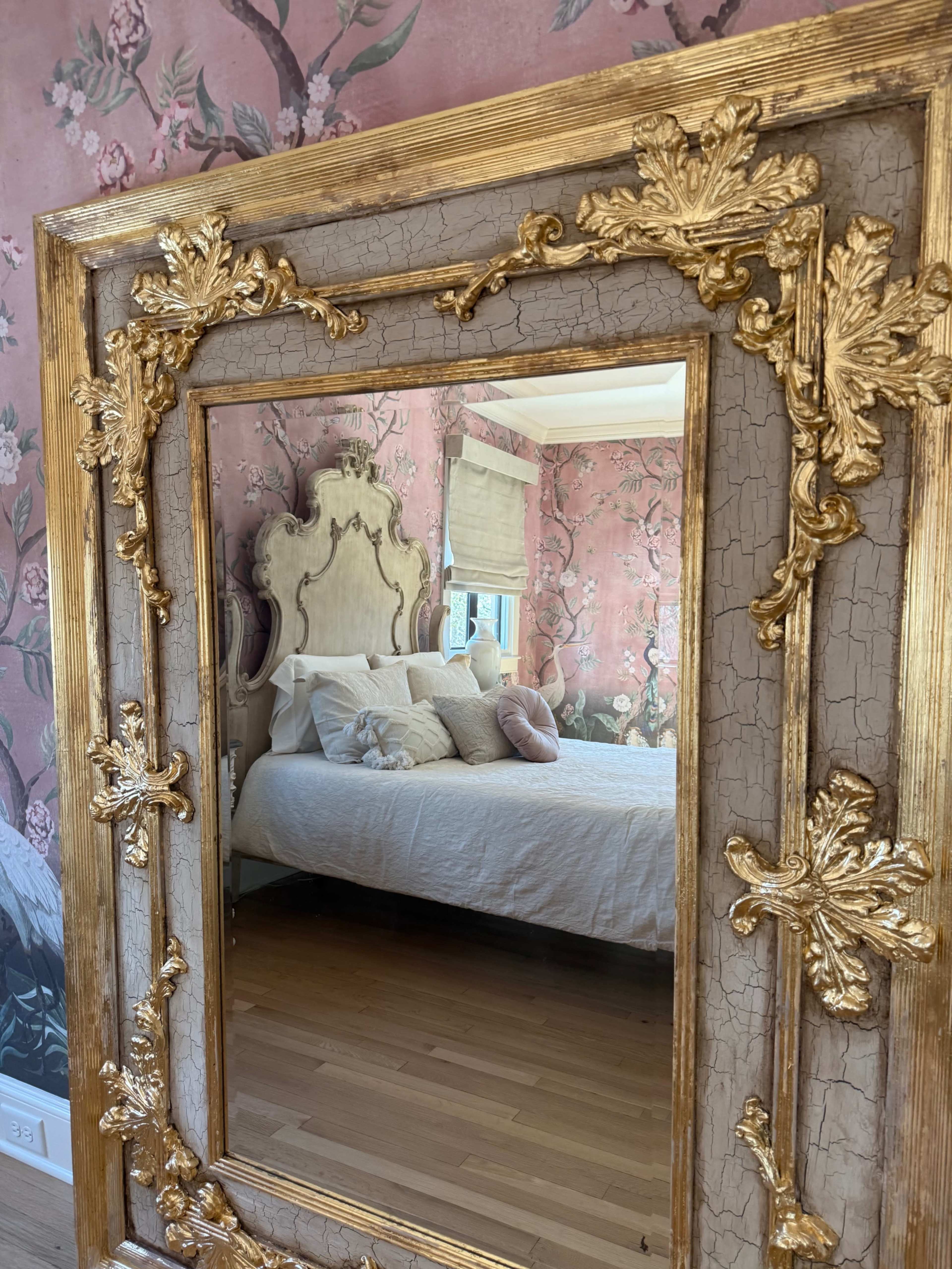 The image shows a decorative gold-framed mirror reflecting a bedroom with a floral wallpaper and a bed adorned with various pillows.