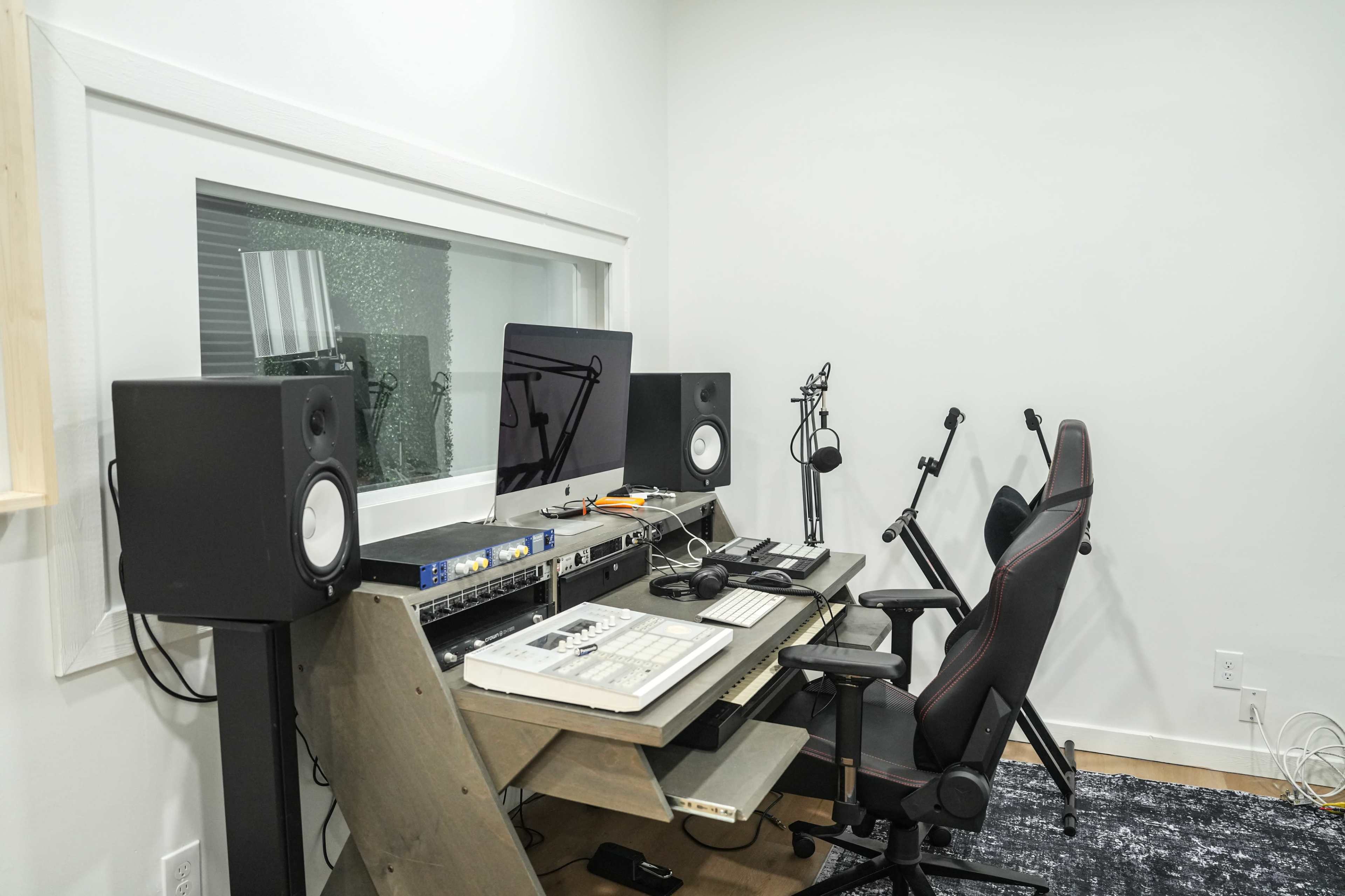 The image shows a modern music studio featuring a computer workstation, audio monitors, and two chairs, with soundproof glass separating it from another room.