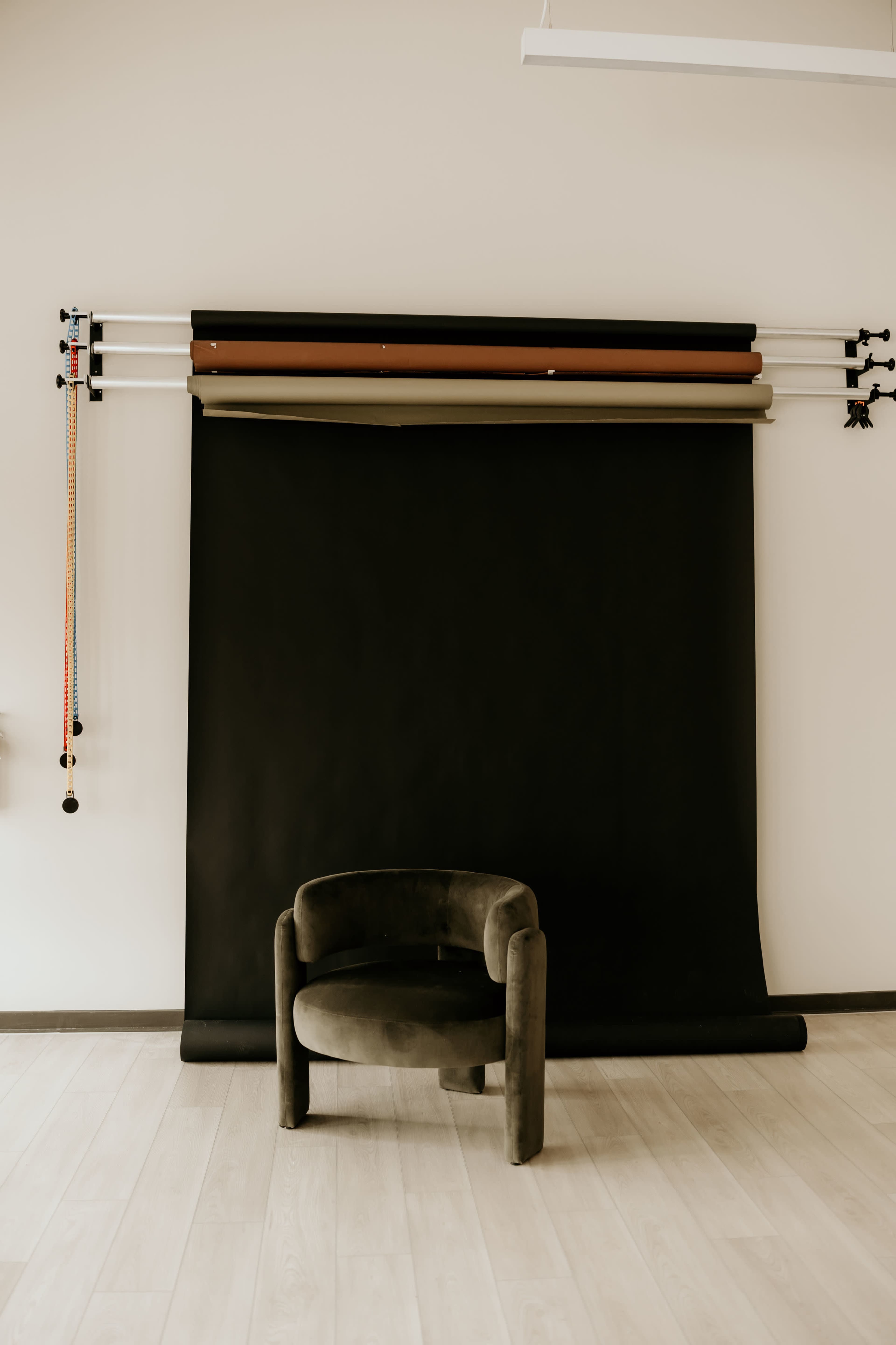 A round, dark upholstered chair is positioned in front of a black photo backdrop mounted on the wall with various colored paper rolls above it.