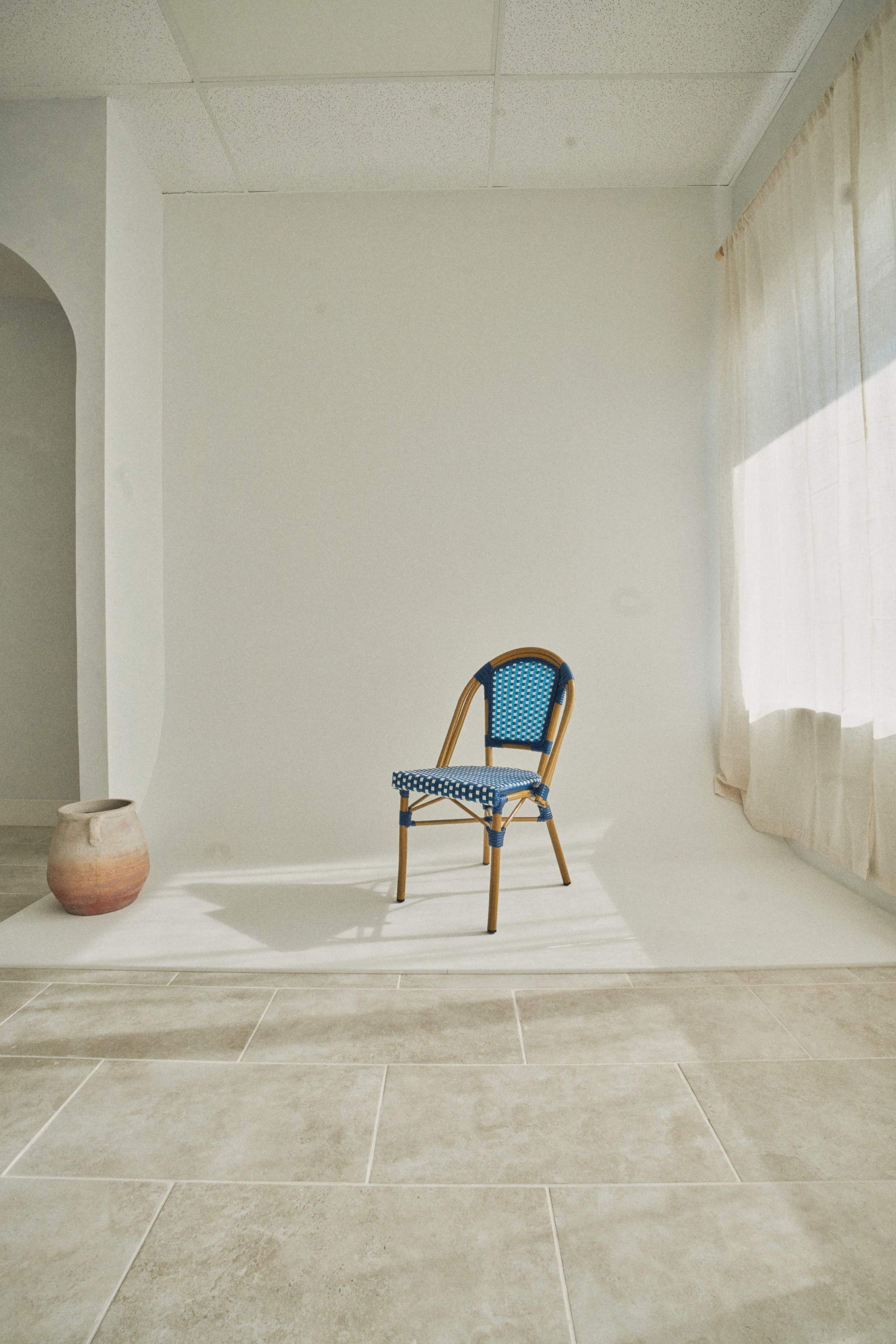 A single wooden chair with a blue seat is positioned in a well-lit room featuring a plain white wall and a clay pot nearby.