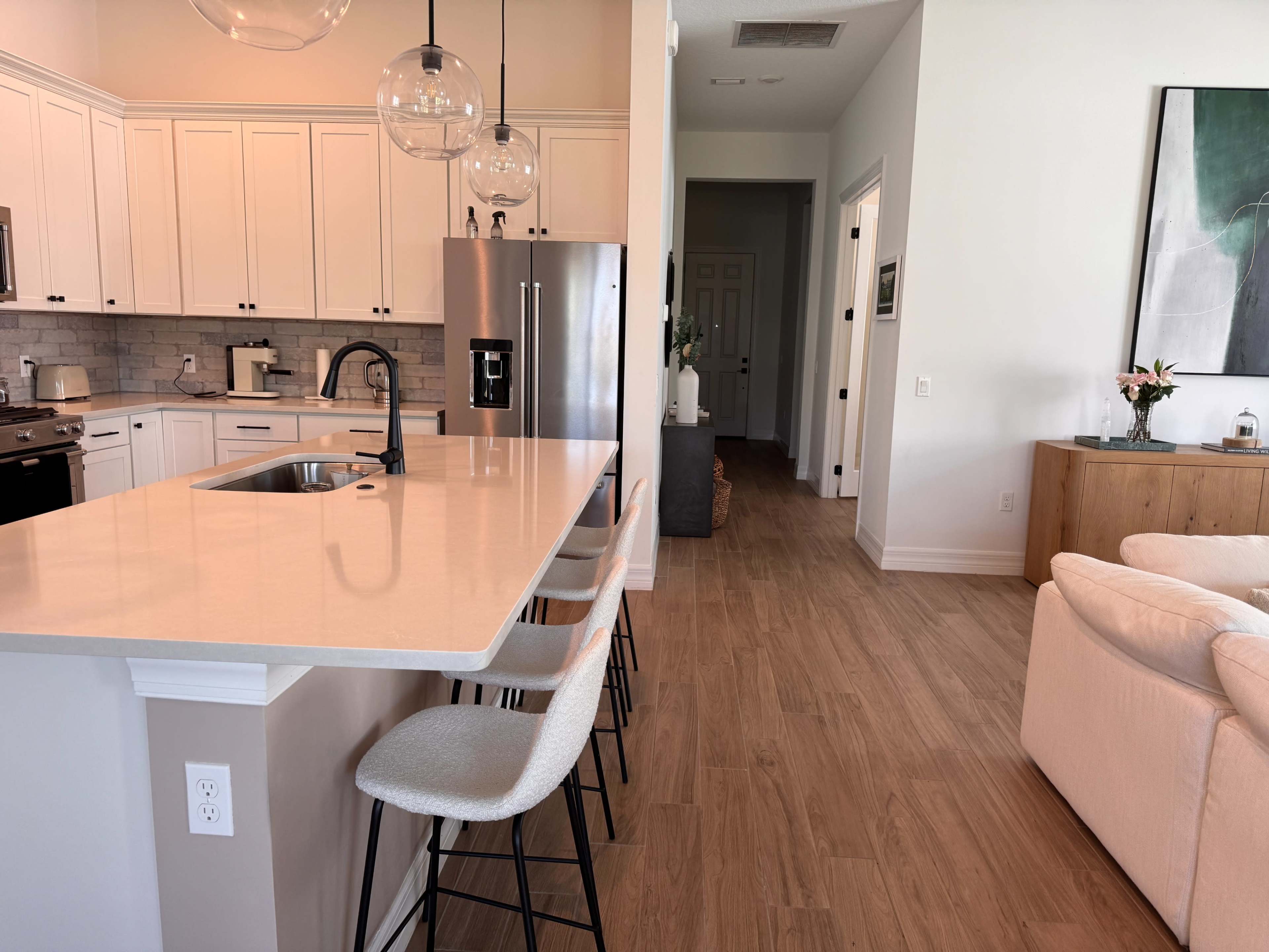 The image shows a modern kitchen with a large island, bar stools, and an open layout leading to a living area.