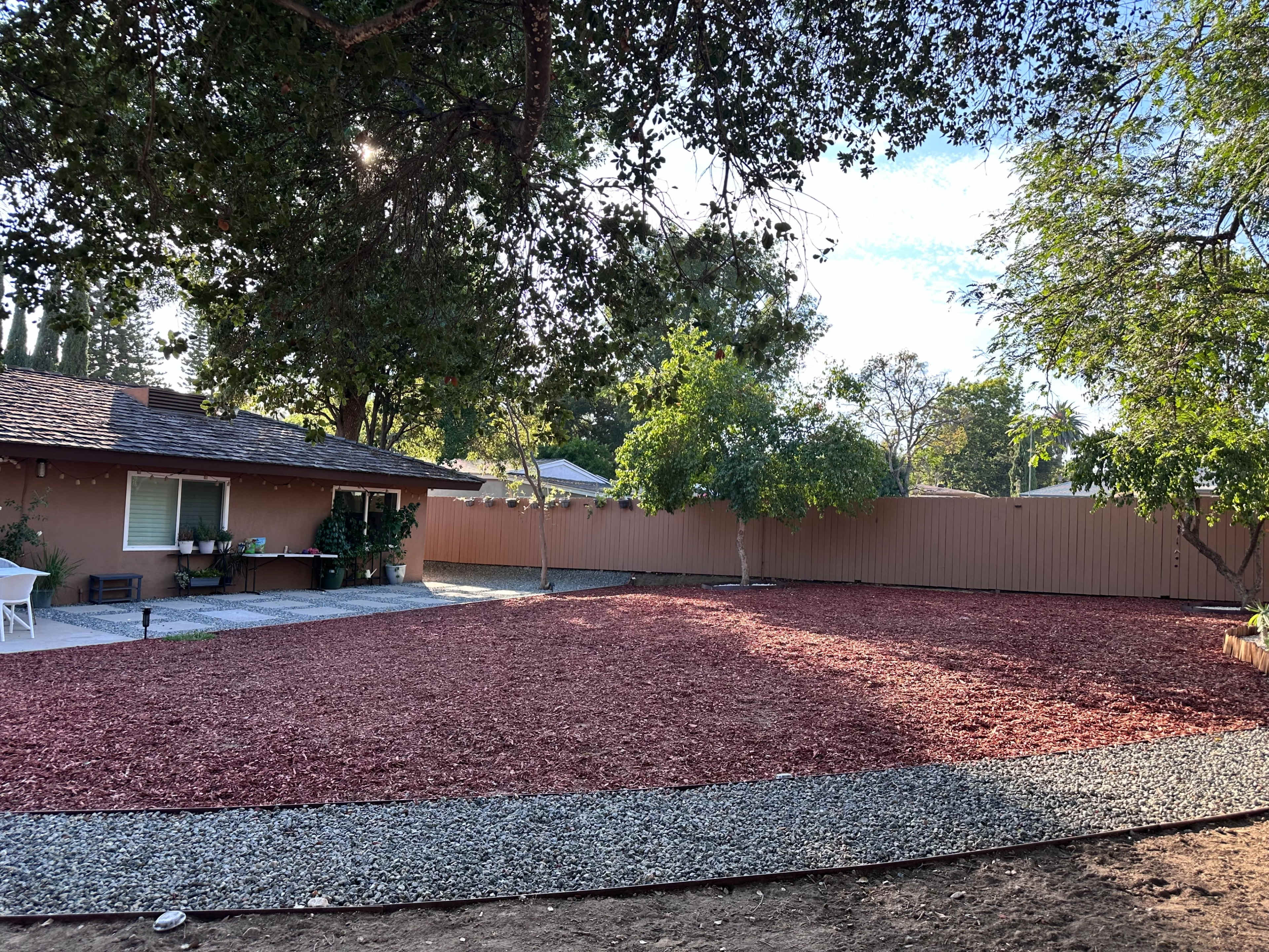 A backyard features a freshly landscaped area with red mulch and a gravel pathway, bordered by a house and trees.