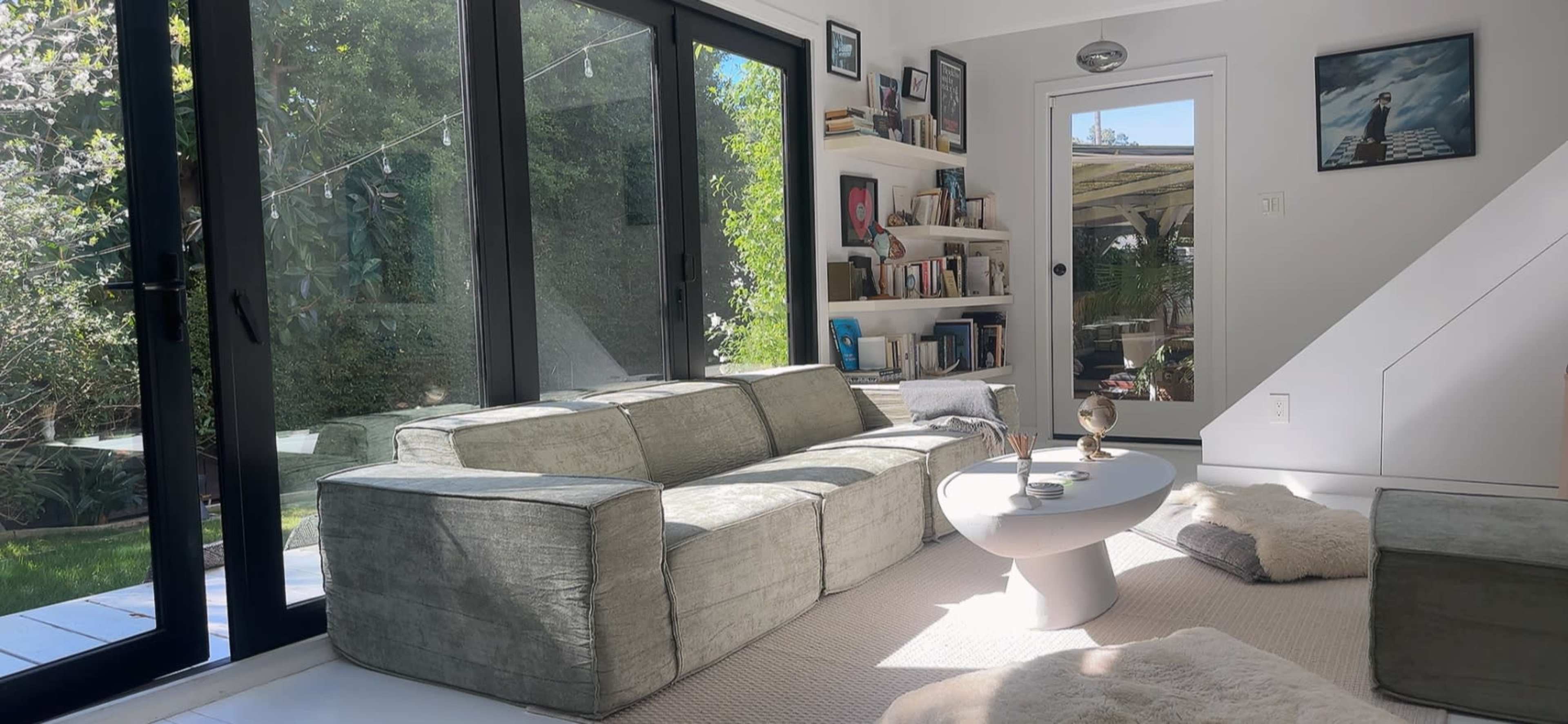 A living room features a gray sofa, a round white coffee table, and bookshelves, with large glass doors opening to a garden outside.