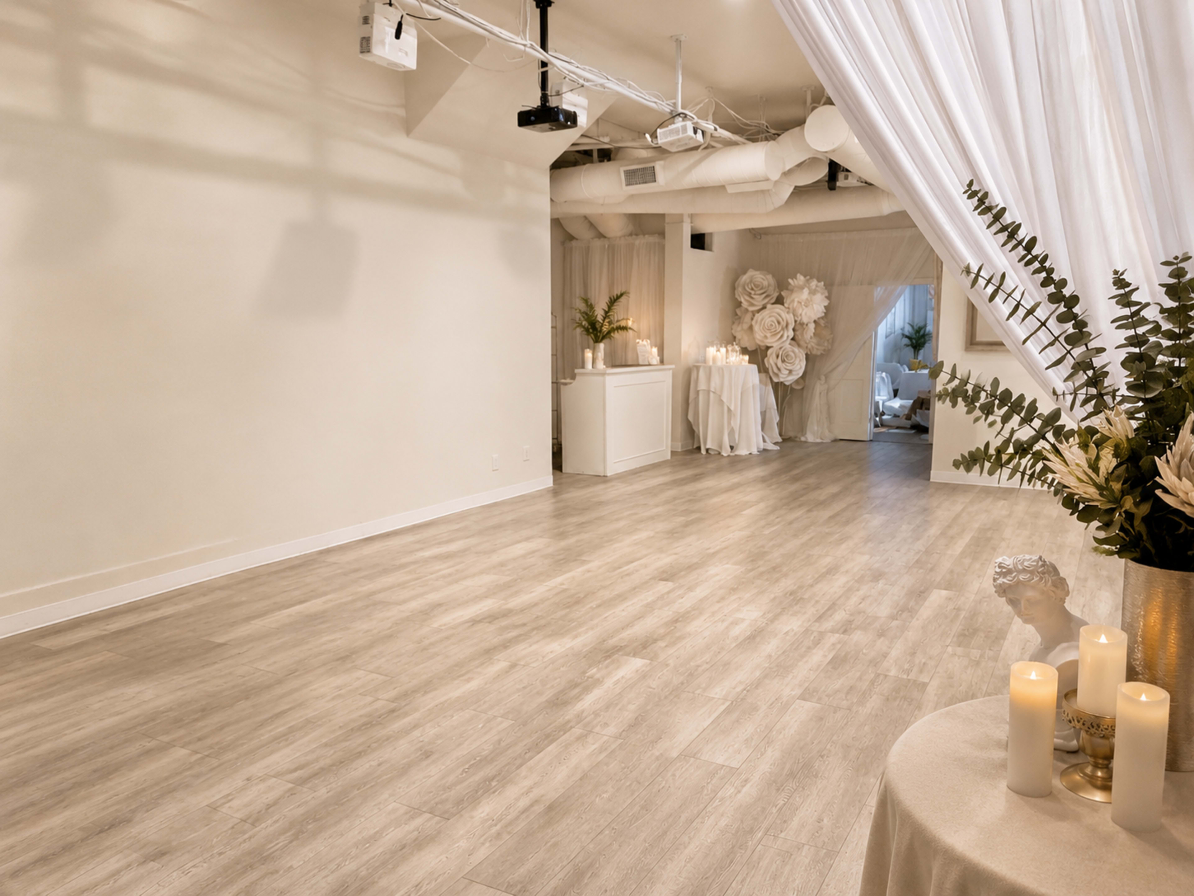 The image shows a spacious, well-lit room with white walls, hardwood flooring, and decorative elements including candles and large floral arrangements.