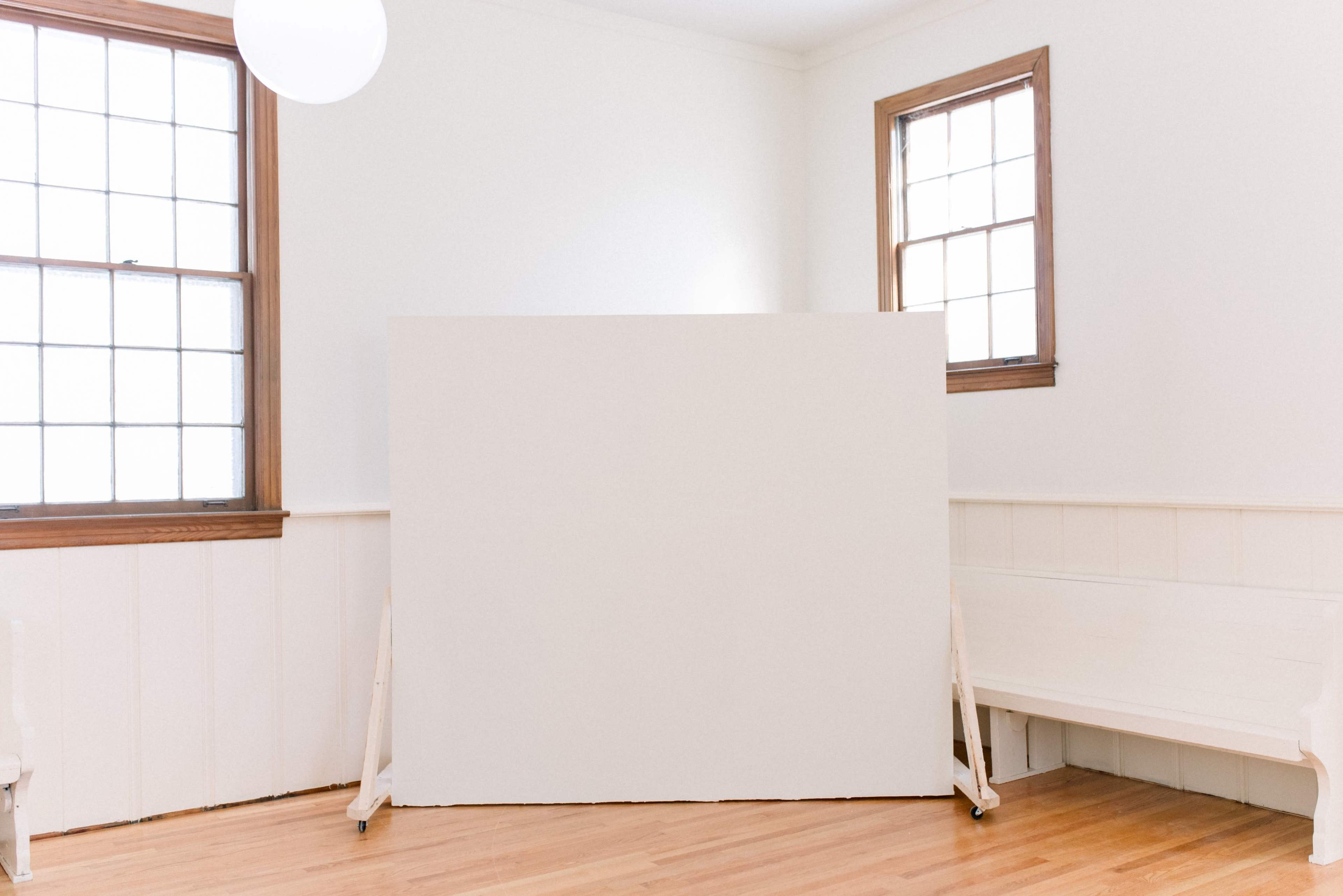 A bright room with white walls, two windows, a blank white canvas on an easel, and a wooden bench along the wall.