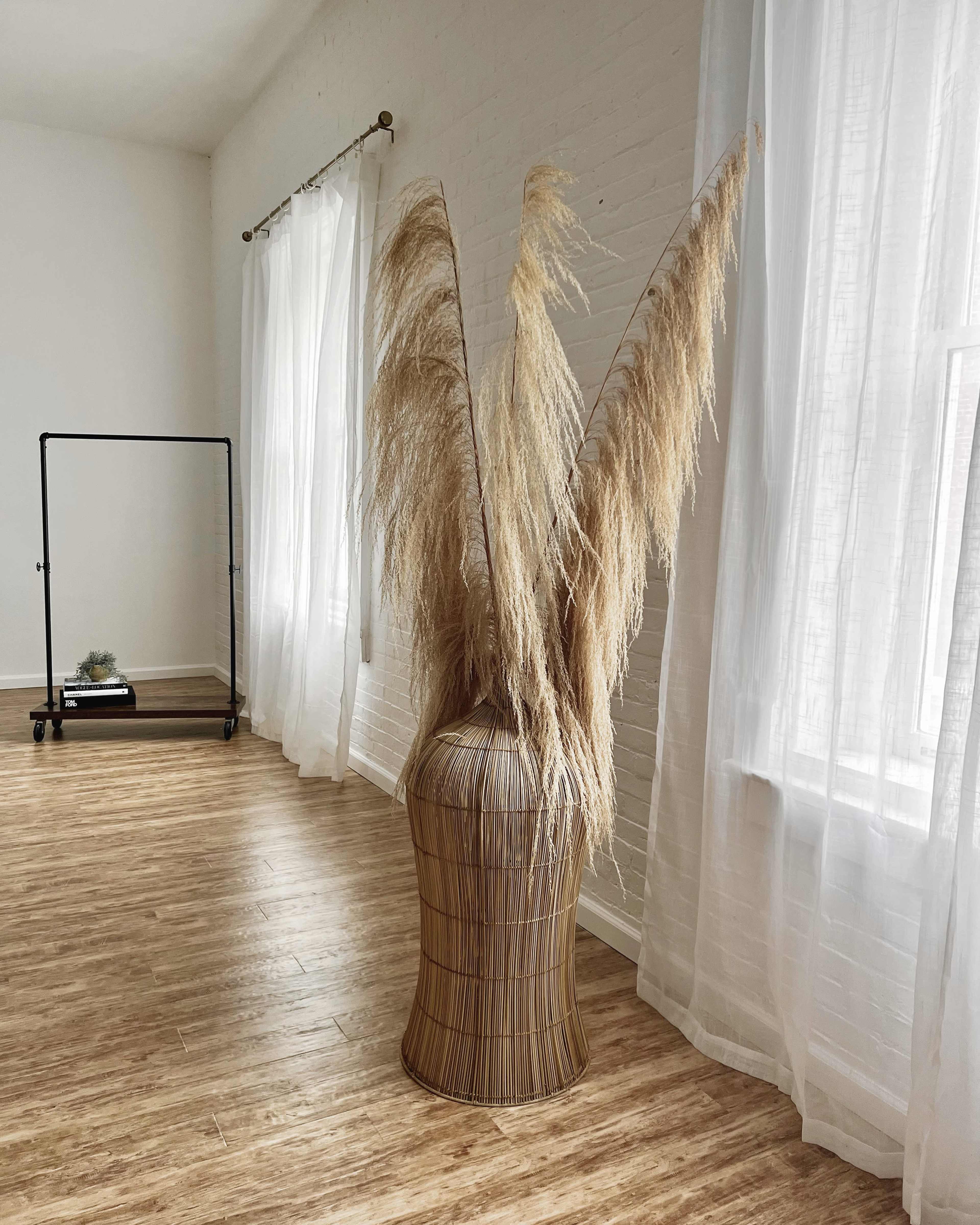A large decorative arrangement of dried pampas grass is placed in a woven basket next to a window with sheer curtains.