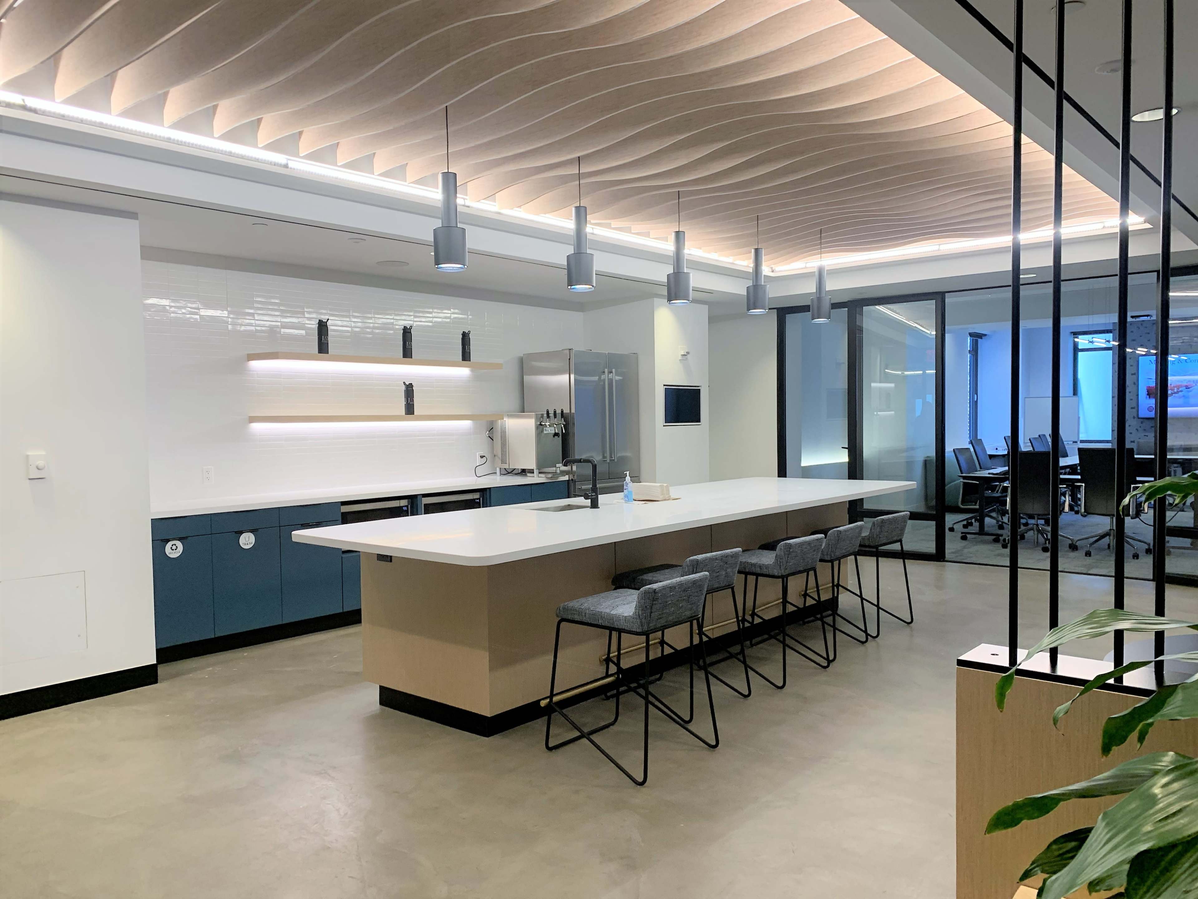The image shows a modern office kitchenette with a large island countertop, bar stools, and a glass-walled meeting room in the background.