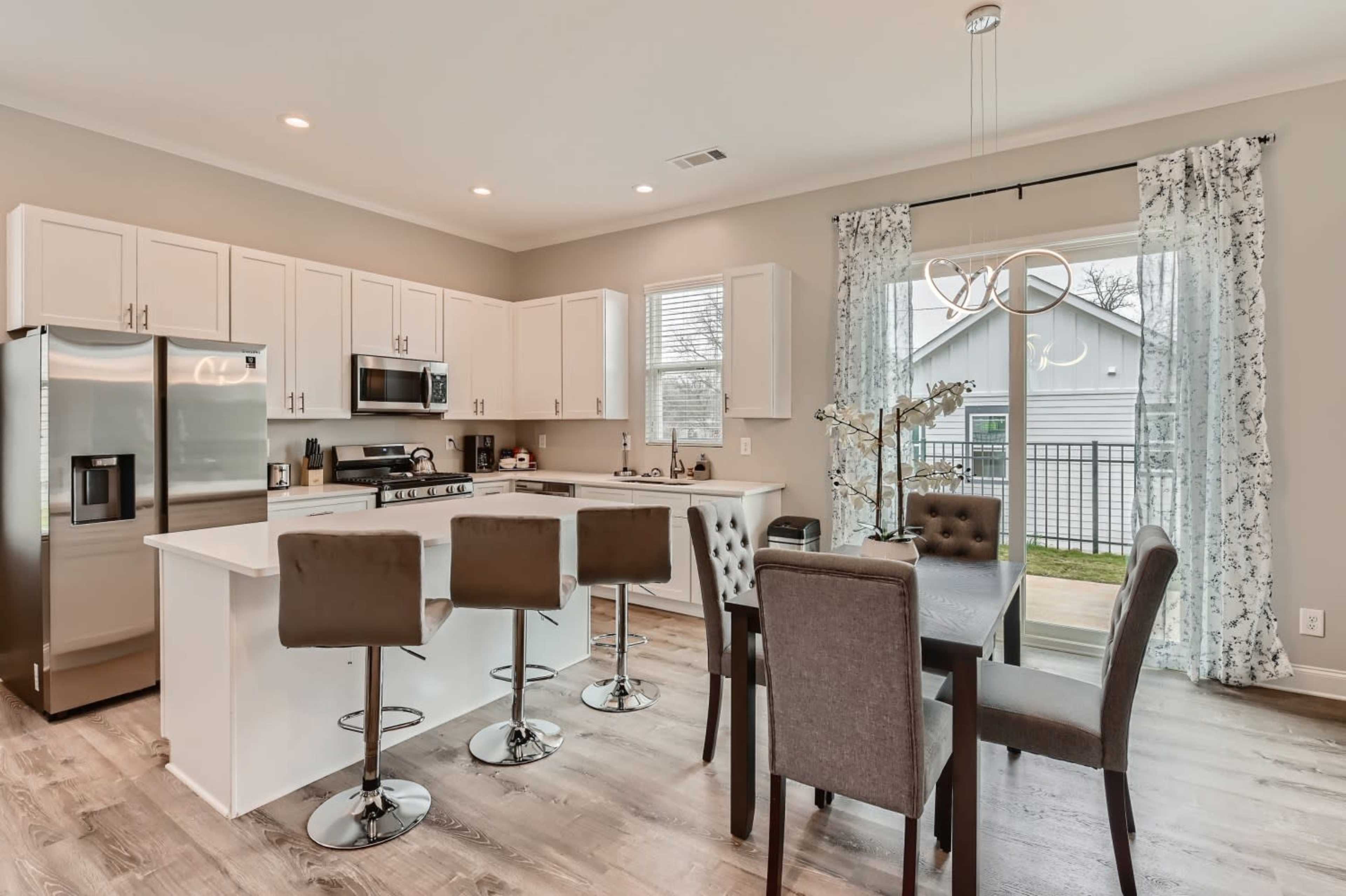 A modern kitchen and dining area with white cabinets, stainless steel appliances, a central island with bar stools, and a dining table set near large windows.