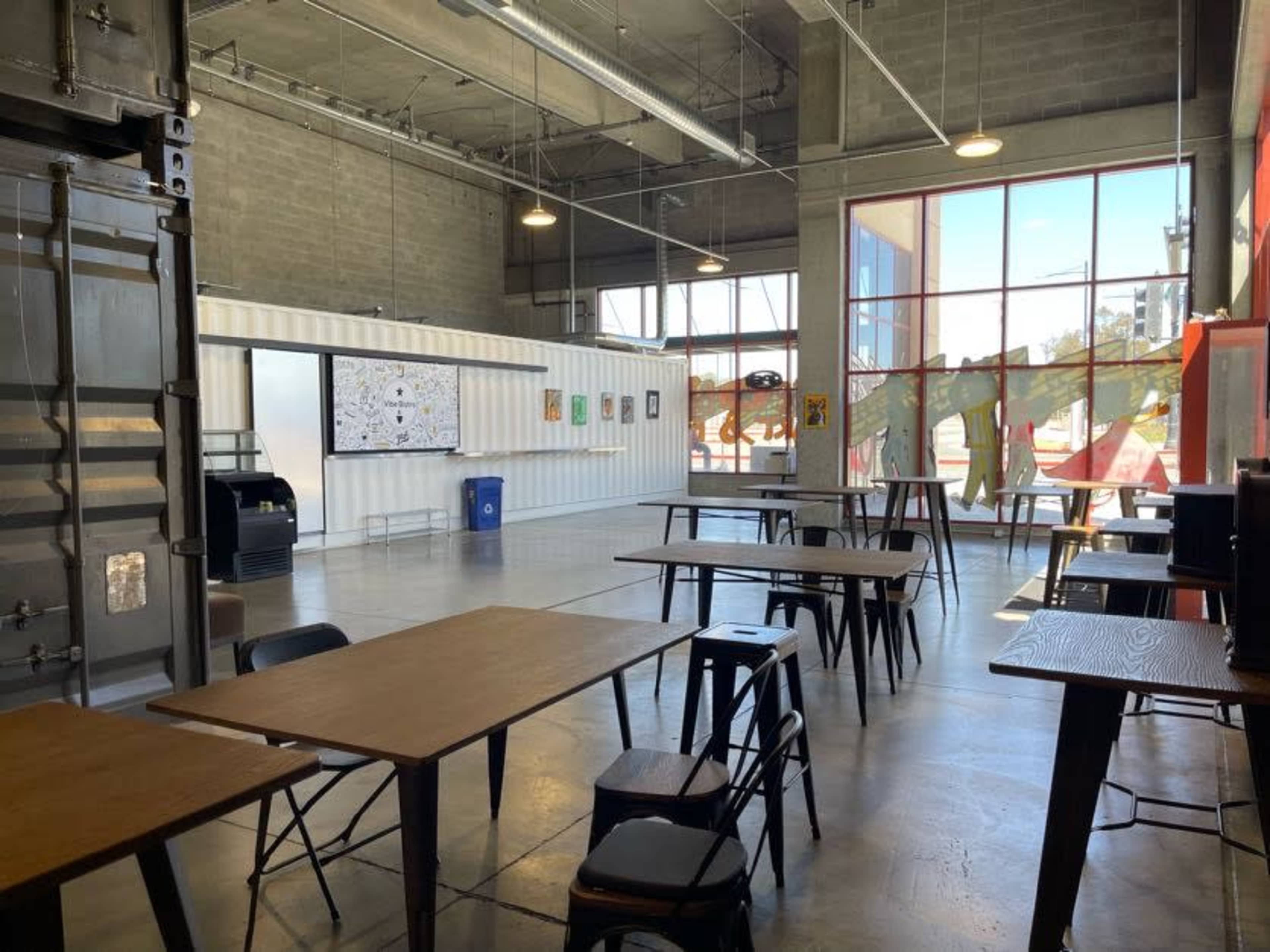 A spacious industrial-style room with several wooden tables and chairs, large windows, and a wall displaying art.