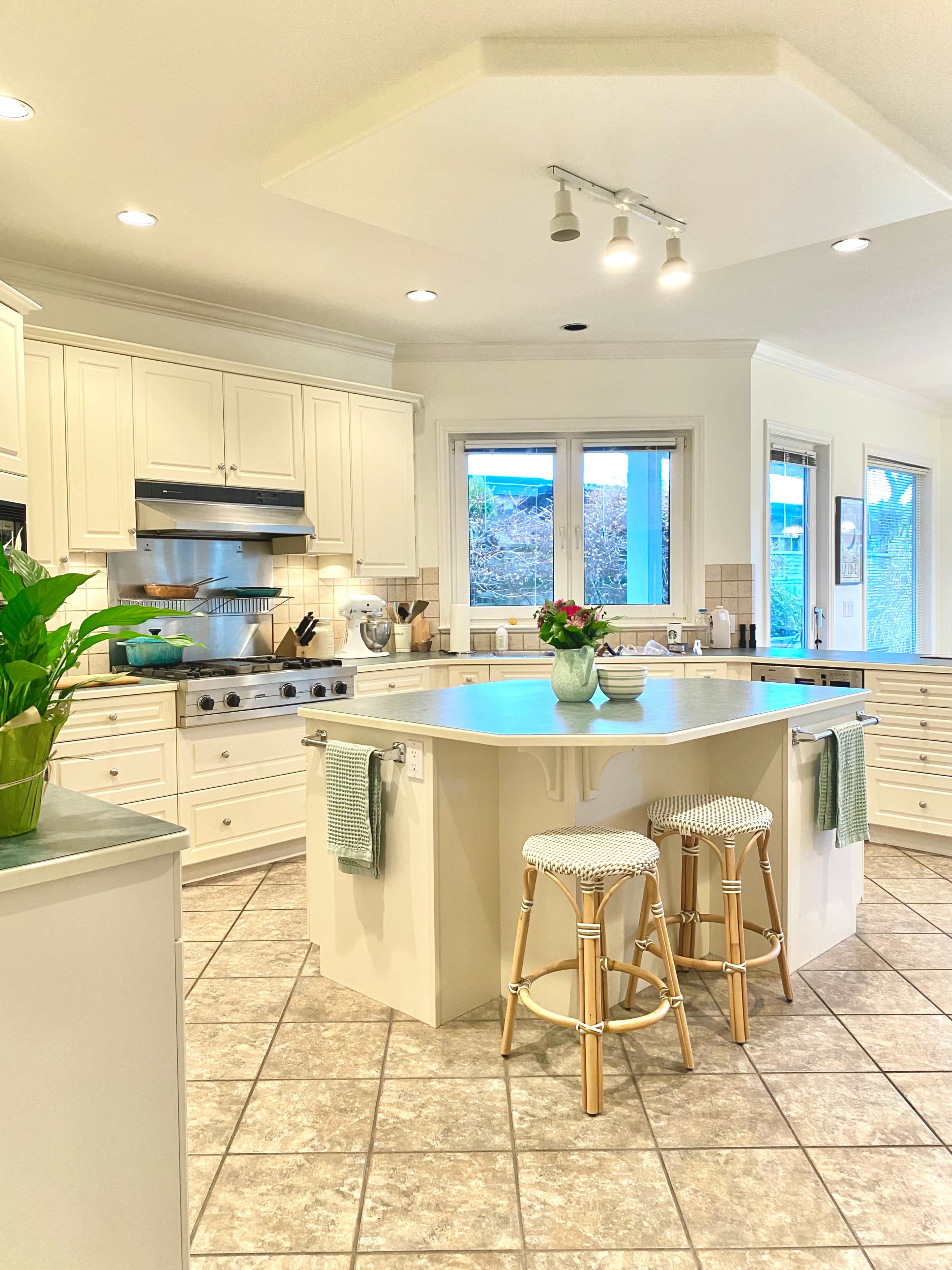 A modern kitchen features light-colored cabinetry, a central island with two stools, and a large window overlooking a garden.