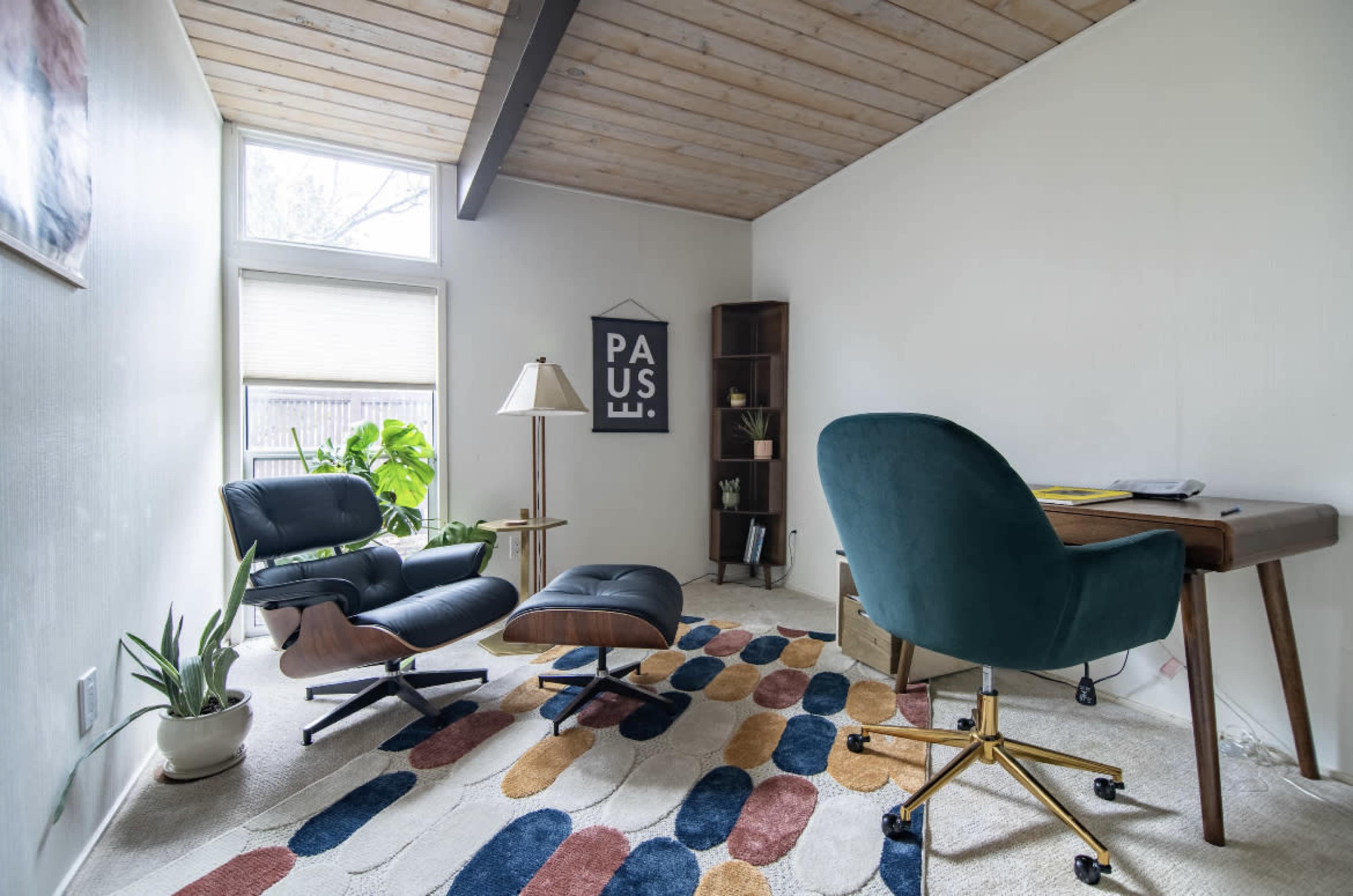 A modern home office with a comfortable chair and ottoman, a desk, plants, and a patterned rug on a carpeted floor.