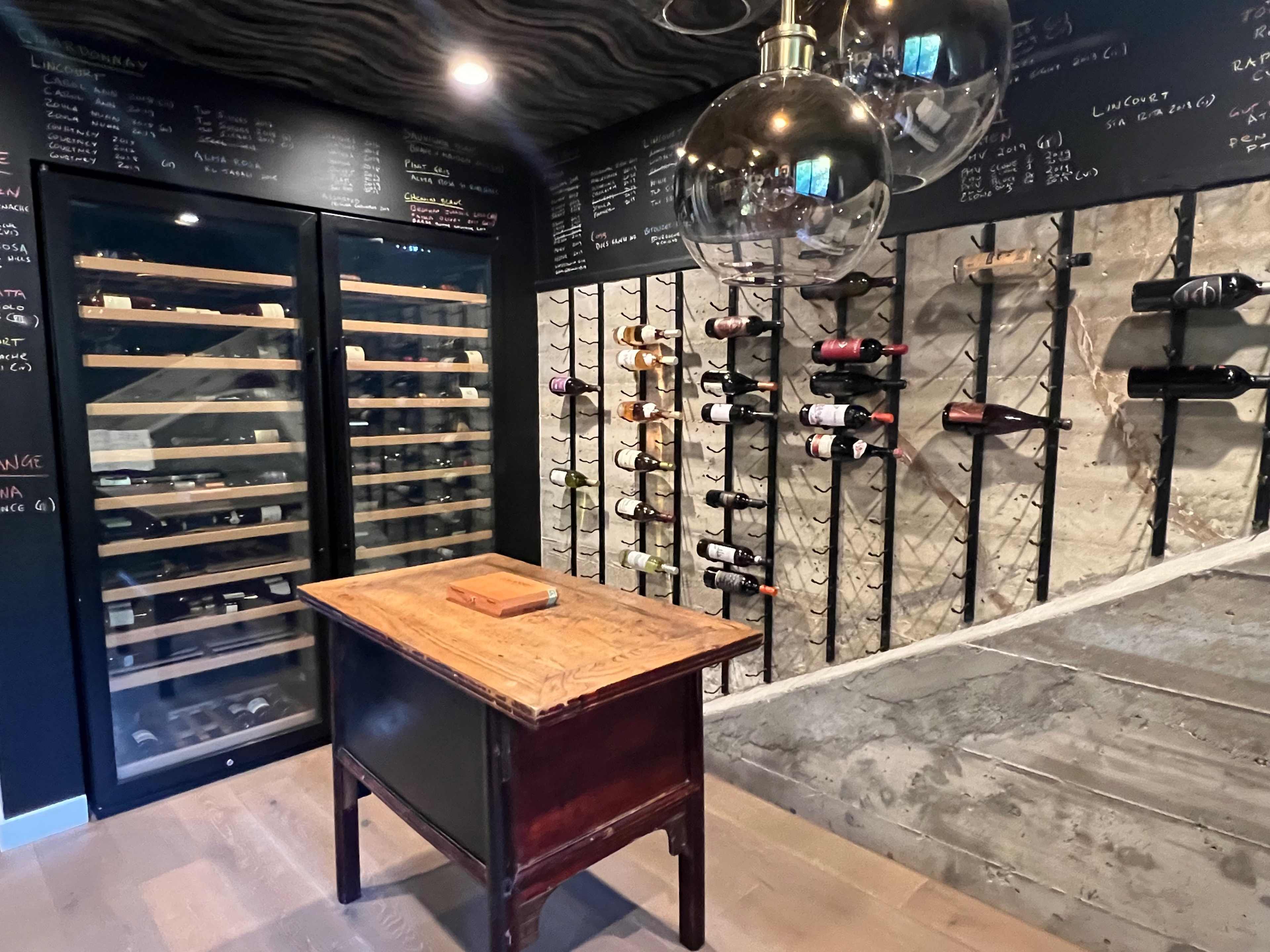 The image shows a wine cellar featuring a glass-fronted wine refrigeration unit on one side, a wall with hanging wine racks, and a wooden table in the center.
